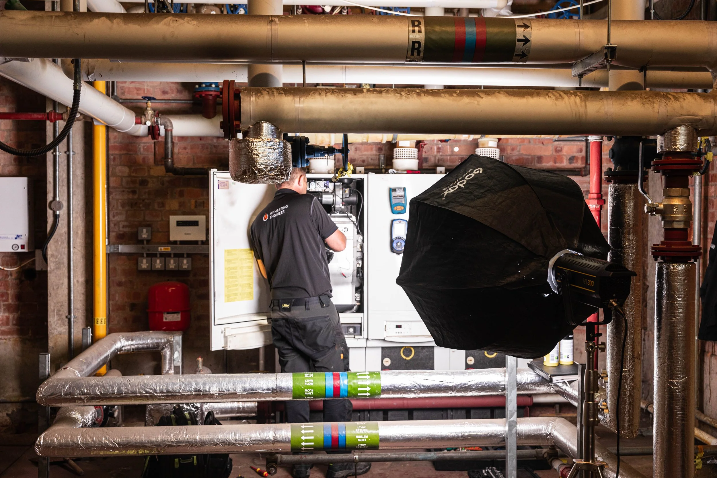 A technician working on a heating system or boiler in an industrial or commercial setting, with pipes and equipment around.