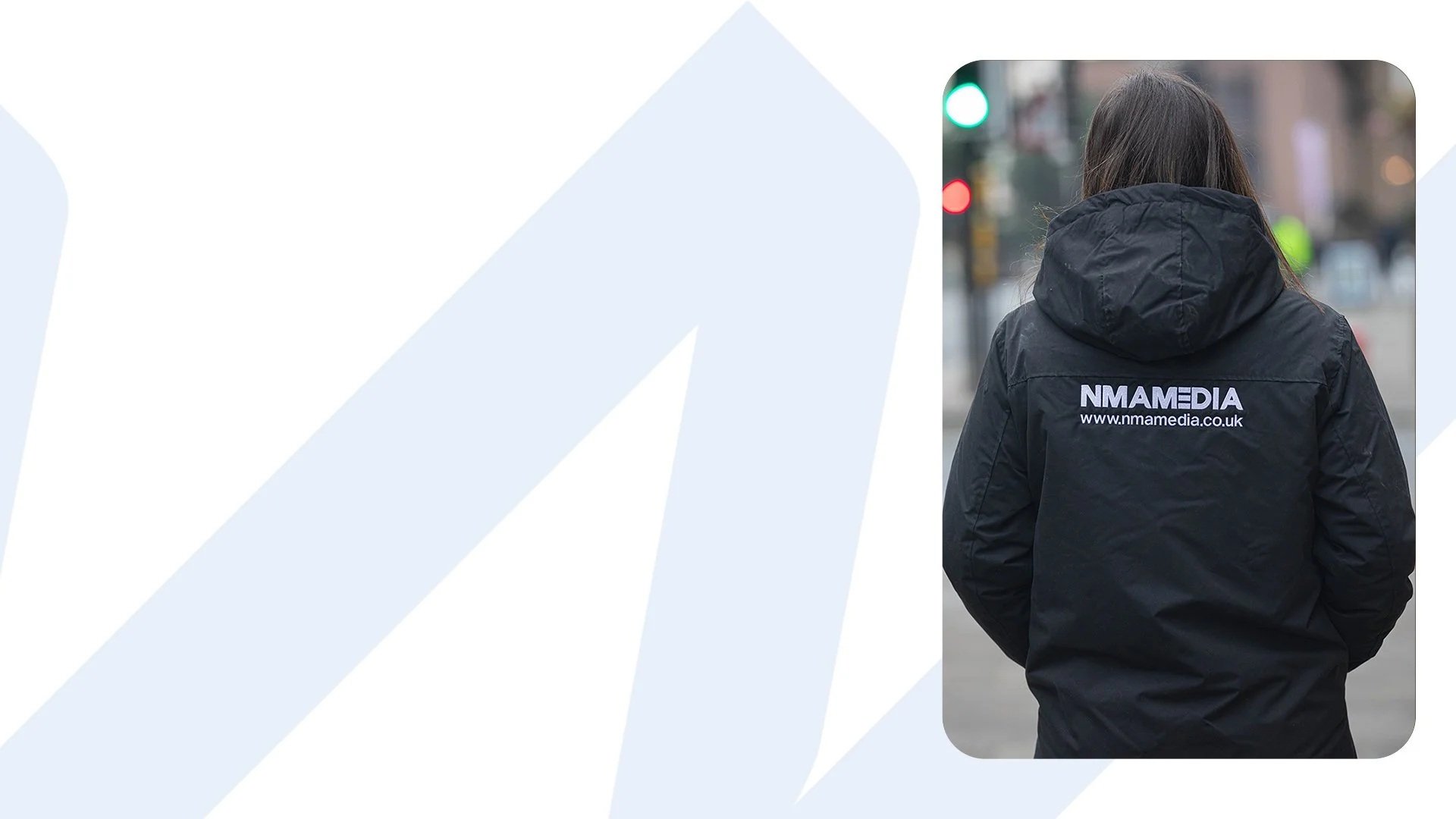 A person with long dark hair wearing a black jacket with the logo and website of NMAMEDIA on the back, standing outdoors in an urban environment with traffic lights and buildings.