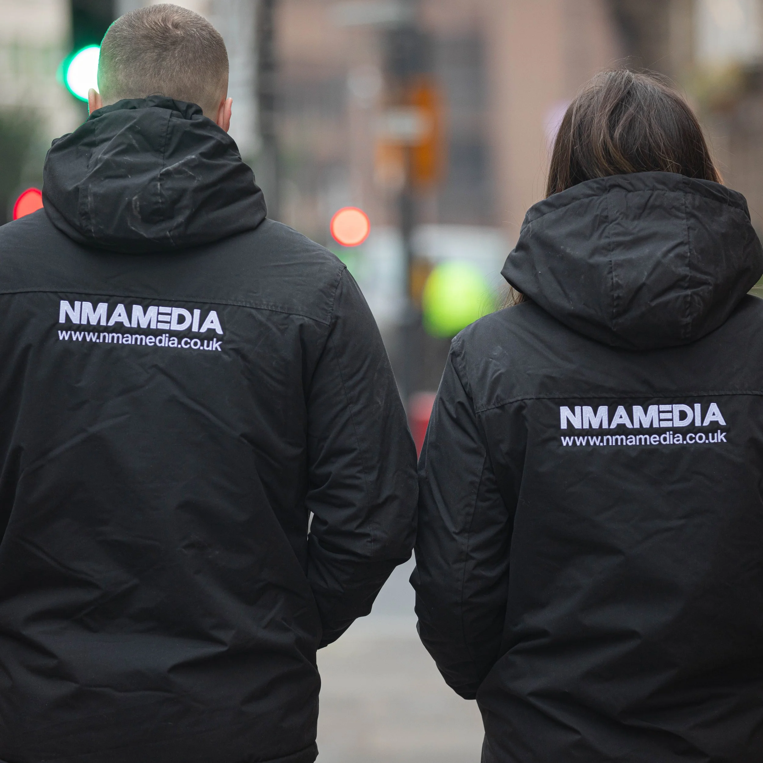 Two people walk together wearing black jackets with the logo and URL 'NMAMEDIA www.nmamedia.co.uk' on the back, with city street blurred background.
