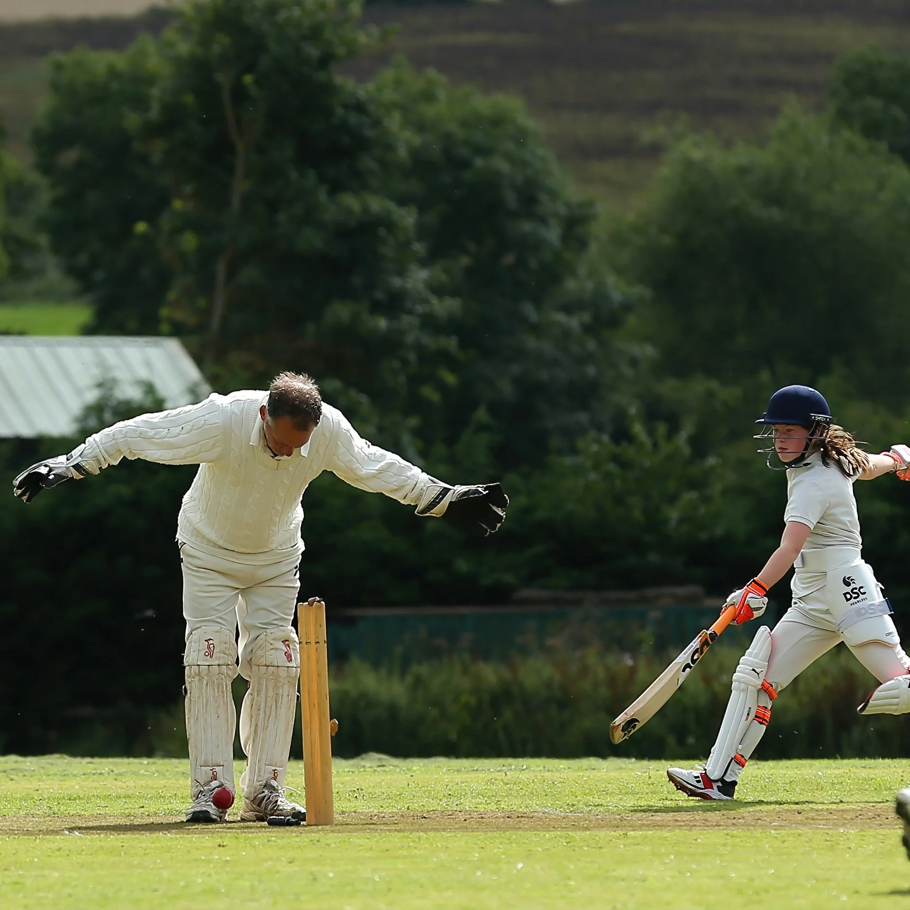 A cricket player with a helmet and pads preparing to hit the ball, while the wicketkeeper in gloves and protective gear watches on.