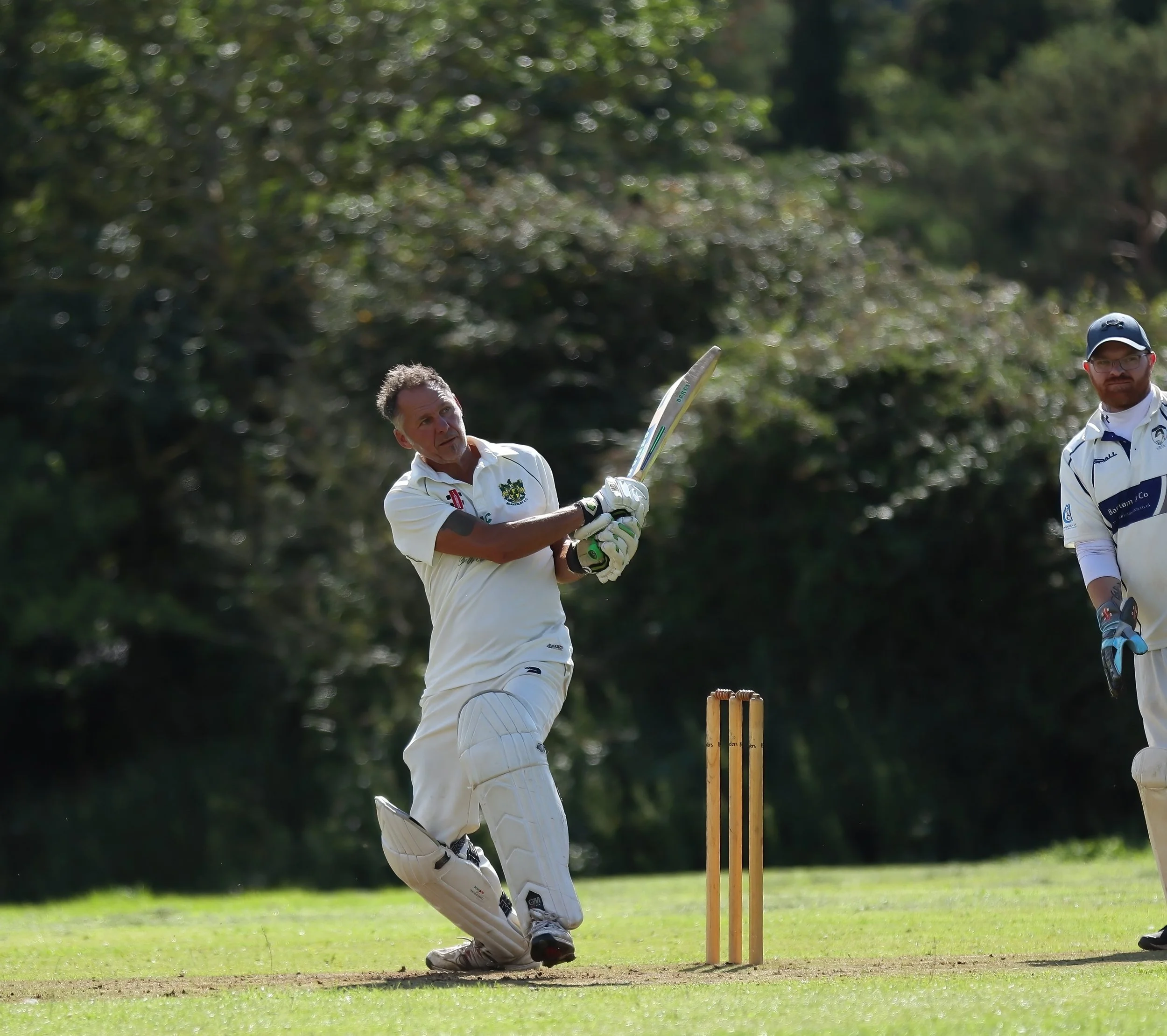 Cricketers playing on the field, with one in a white uniform batting near the stumps while another in a white uniform and cap standing nearby.