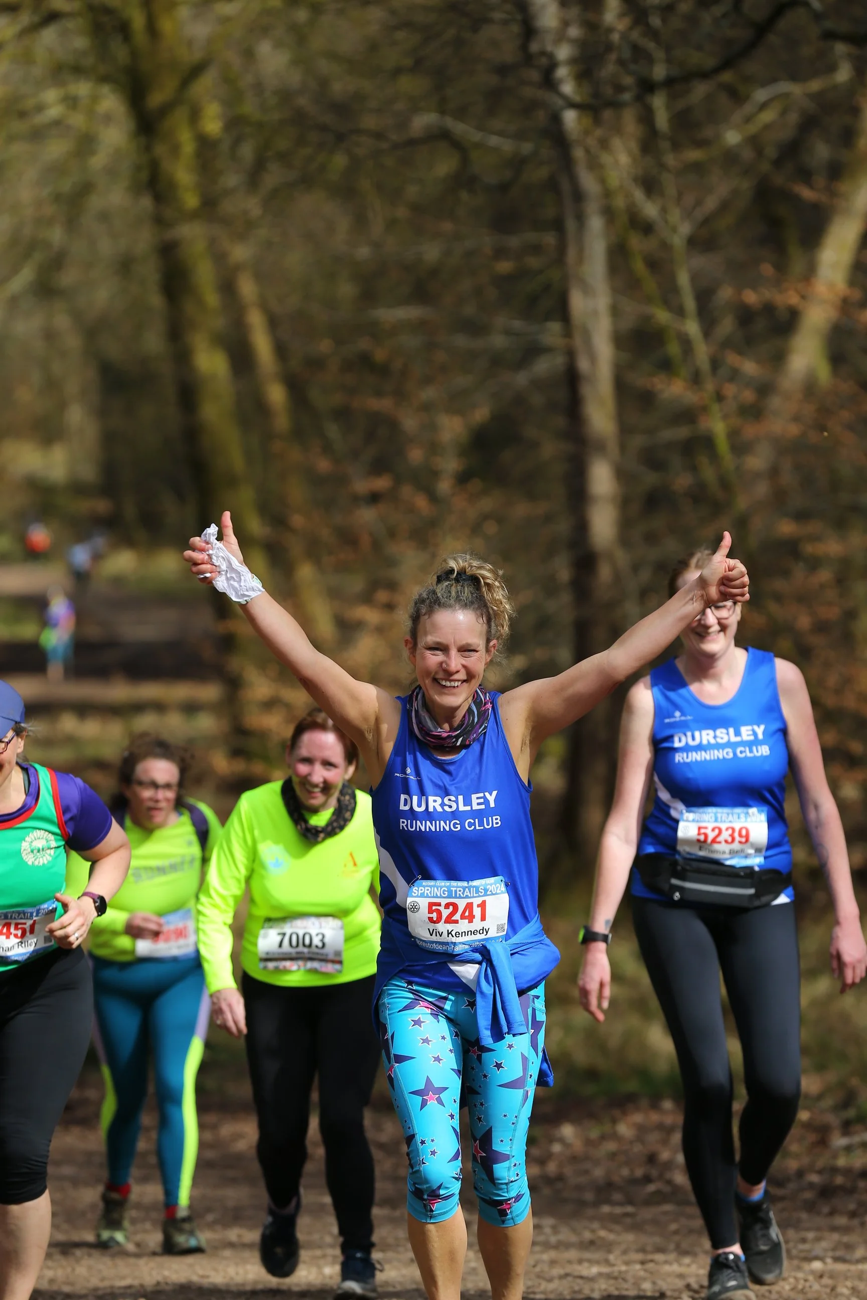 Group of women participating in an outdoor running event in a wooded area, with the woman in the foreground smiling, raising her arms in celebration, and wearing a blue running top and star-patterned leggings.