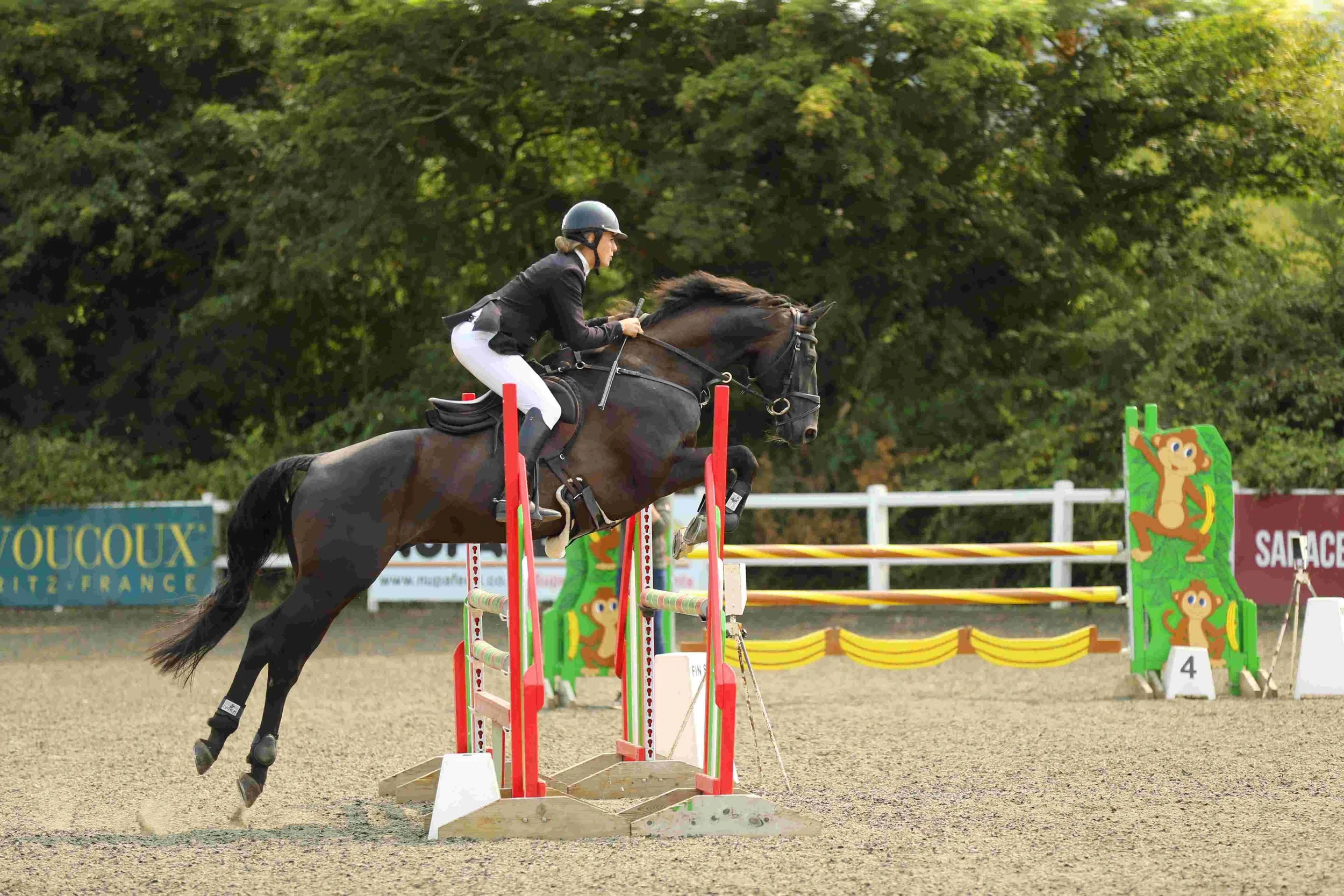 A female equestrian in a black riding jacket, white breeches, and a black helmet jumps over a red and white obstacle with a cartoon monkey on it during an outdoor horse jumping competition, with trees in the background.