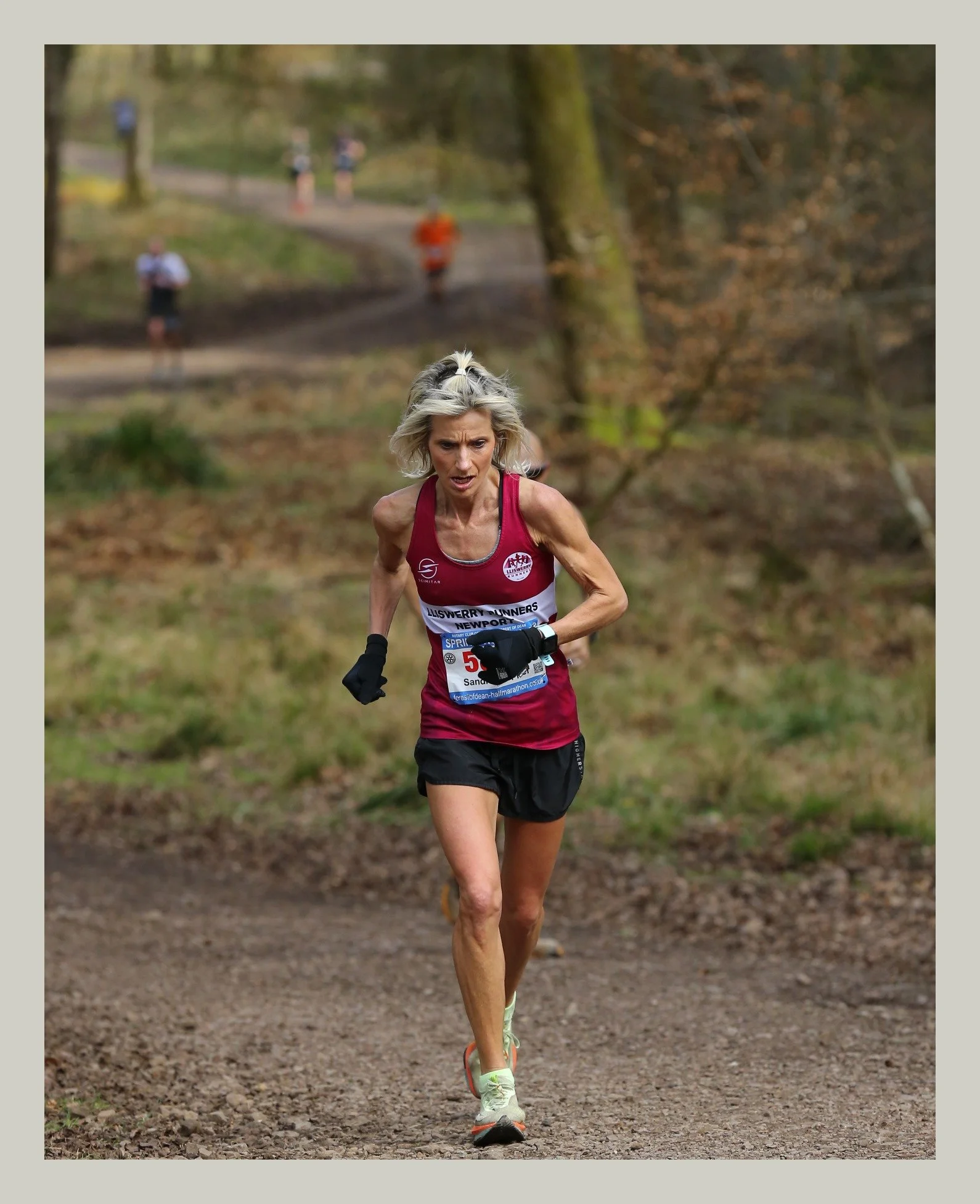 A female runner with blonde hair, wearing a red tank top, black shorts, and white running shoes, running on a forest trail with trees and other runners in the background.