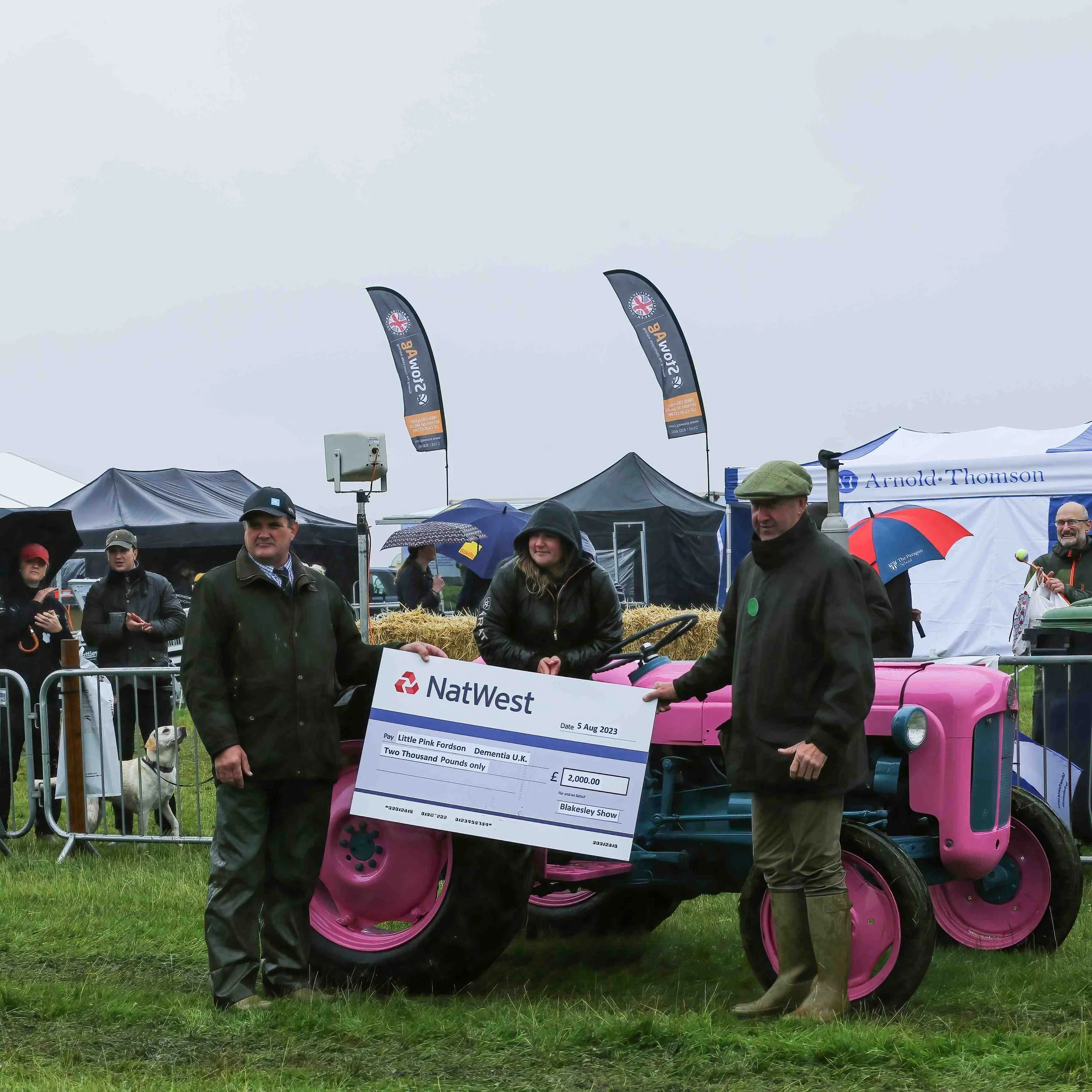People holding a large check next to a pink tractor at the Blakesley Show during a rainy day, with tents and flags in the background.
