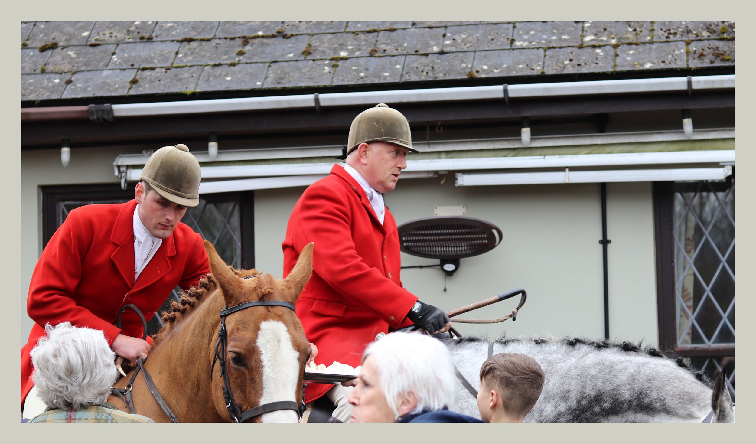 Two men in red coats and helmets riding horses among a crowd outside a building with diamond-patterned windows.