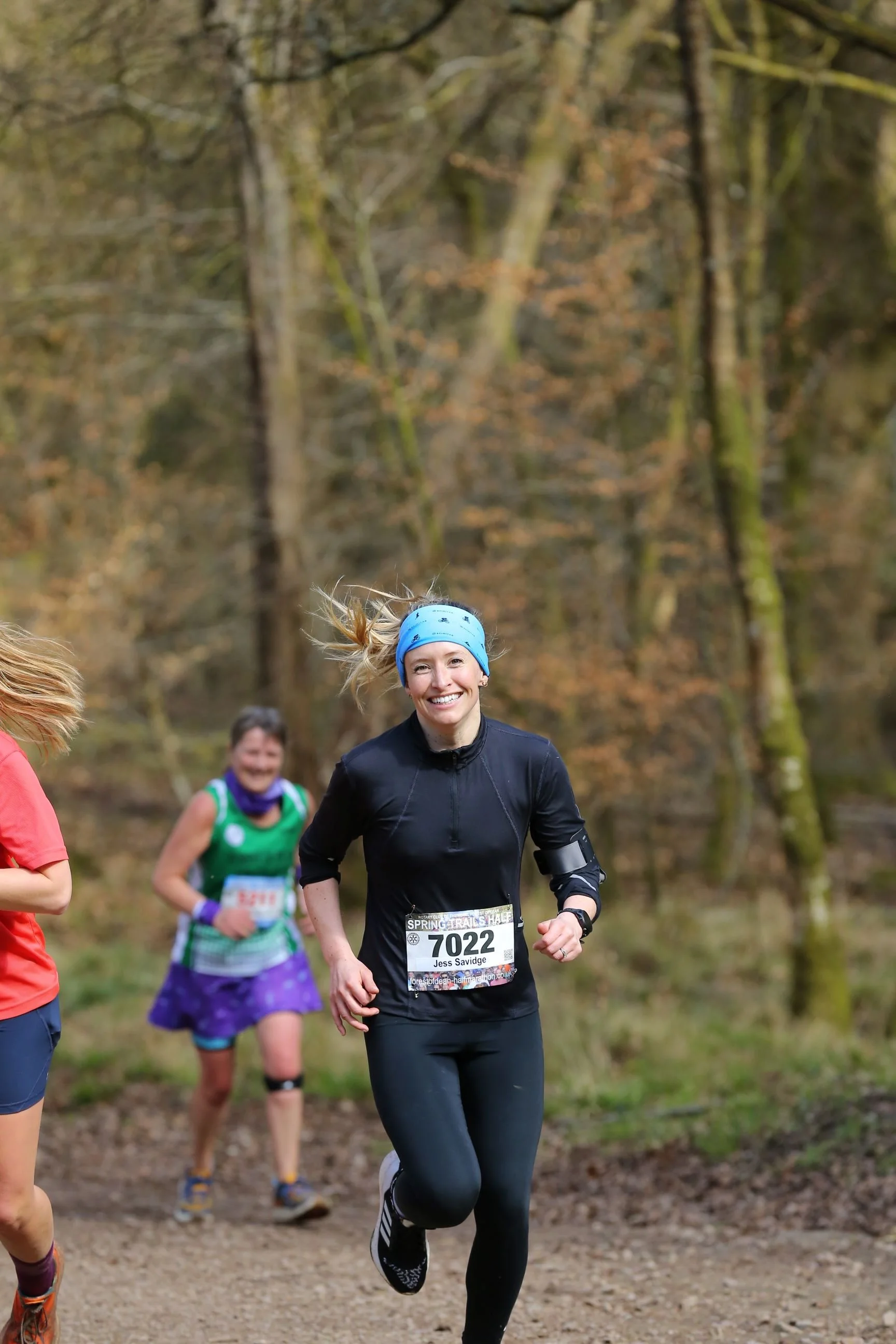 Women running in a forest during a race, smiling and wearing athletic gear with race bibs.
