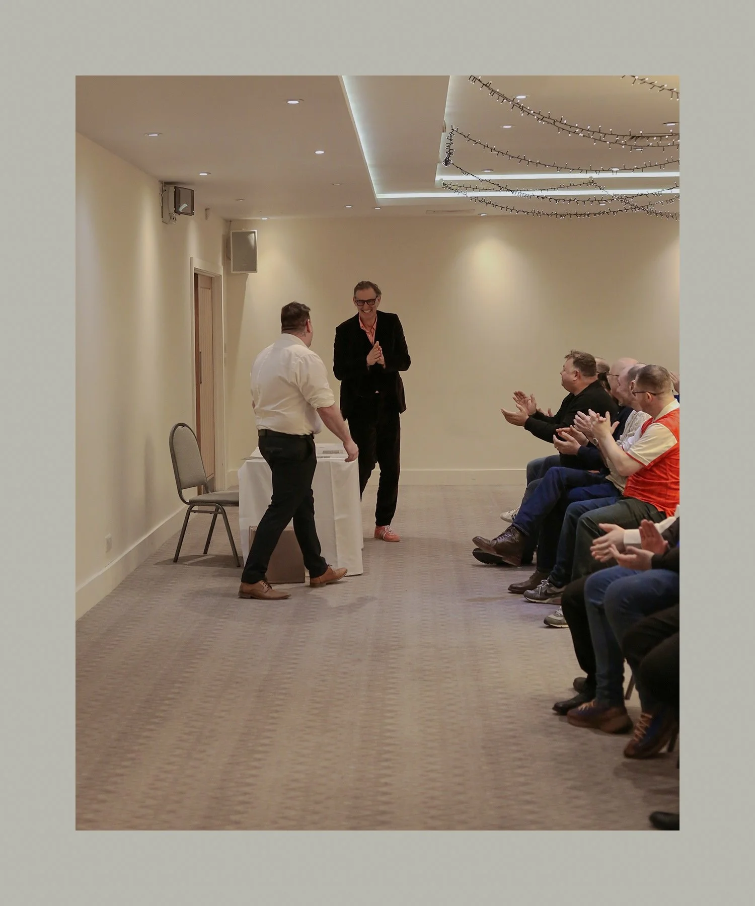 A man in white shirt receiving an award or recognition from a woman in a black suit while an audience claps and smiles in a room decorated with string lights.