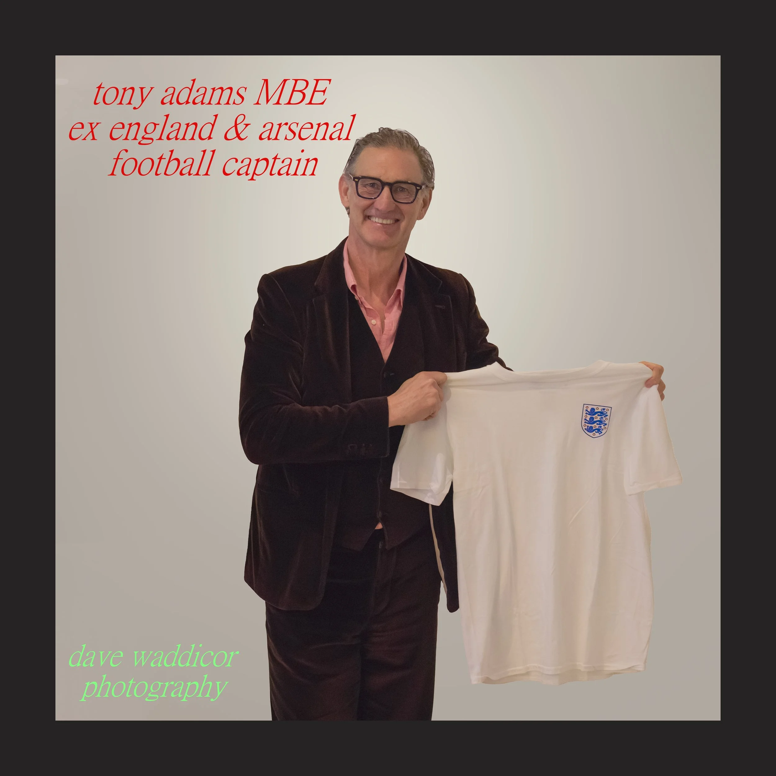 A man in a dark blazer and pink shirt holding a white England football jersey with a blue crest, smiling at camera, against plain light background, with red and green text overlay about his football career and photography credit.