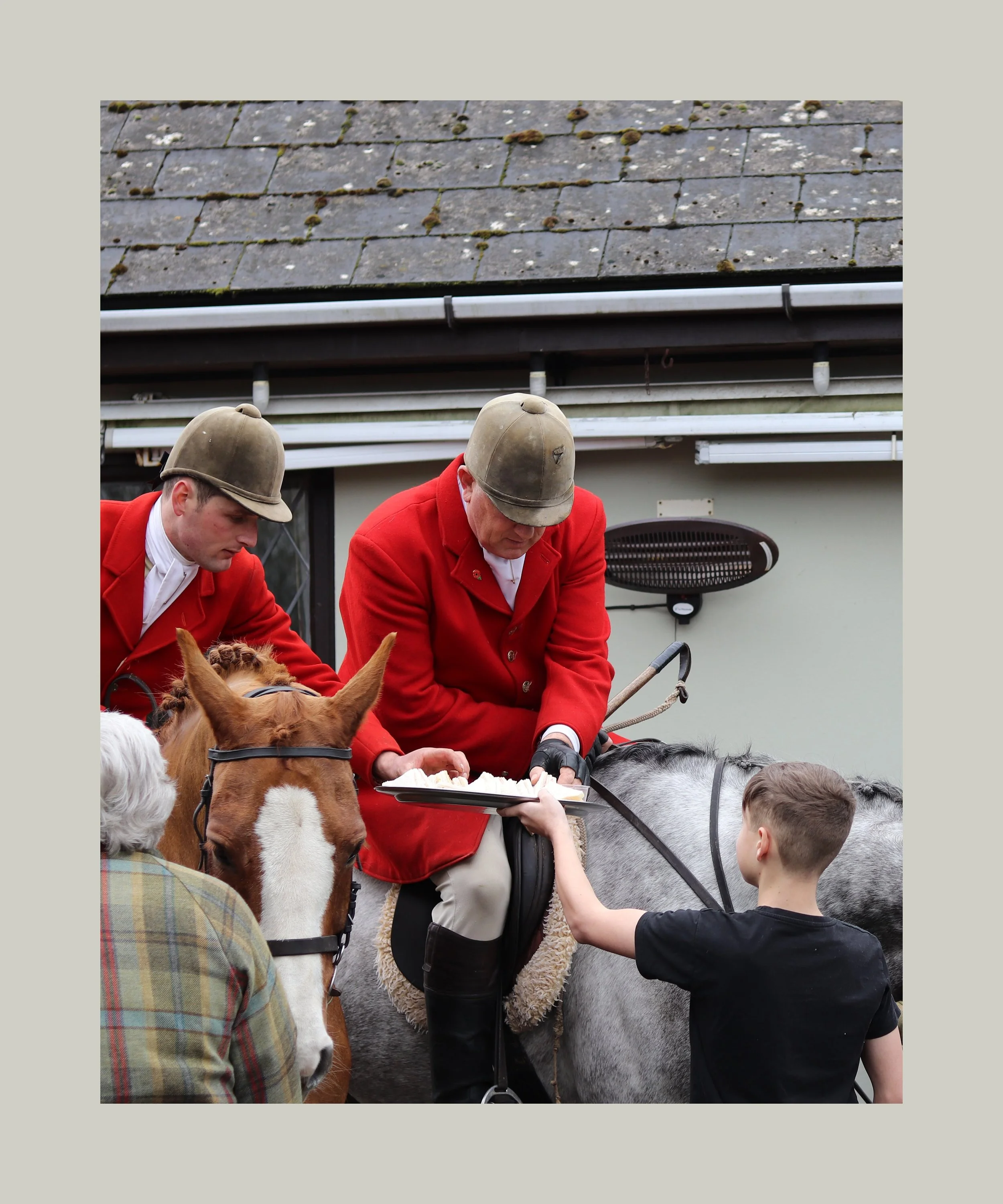 Two men in red jackets and helmets are riding horses. A young boy is offering something to one of the men on the horse. An older woman with white hair is standing nearby.