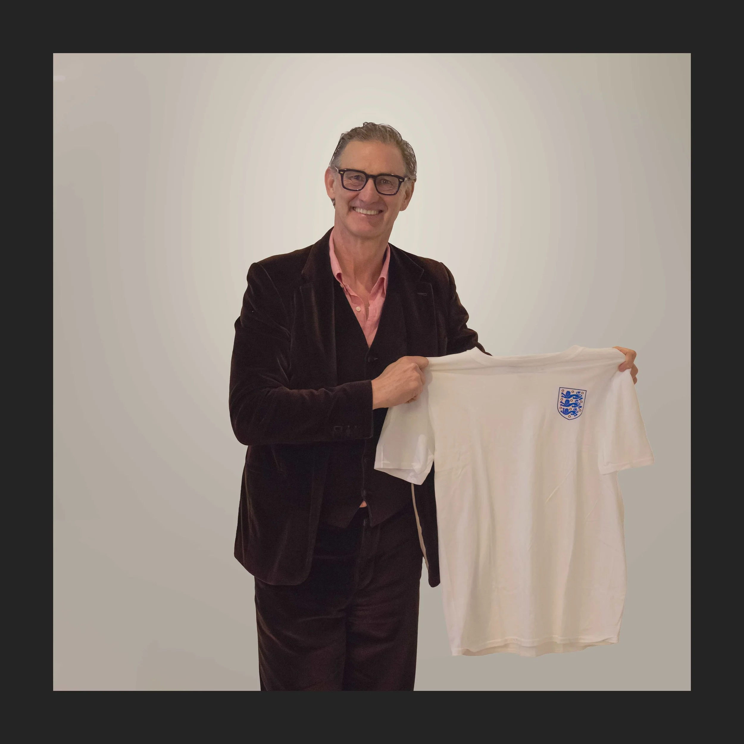 A man in glasses and a brown blazer holding an England football shirt with a crest on it.