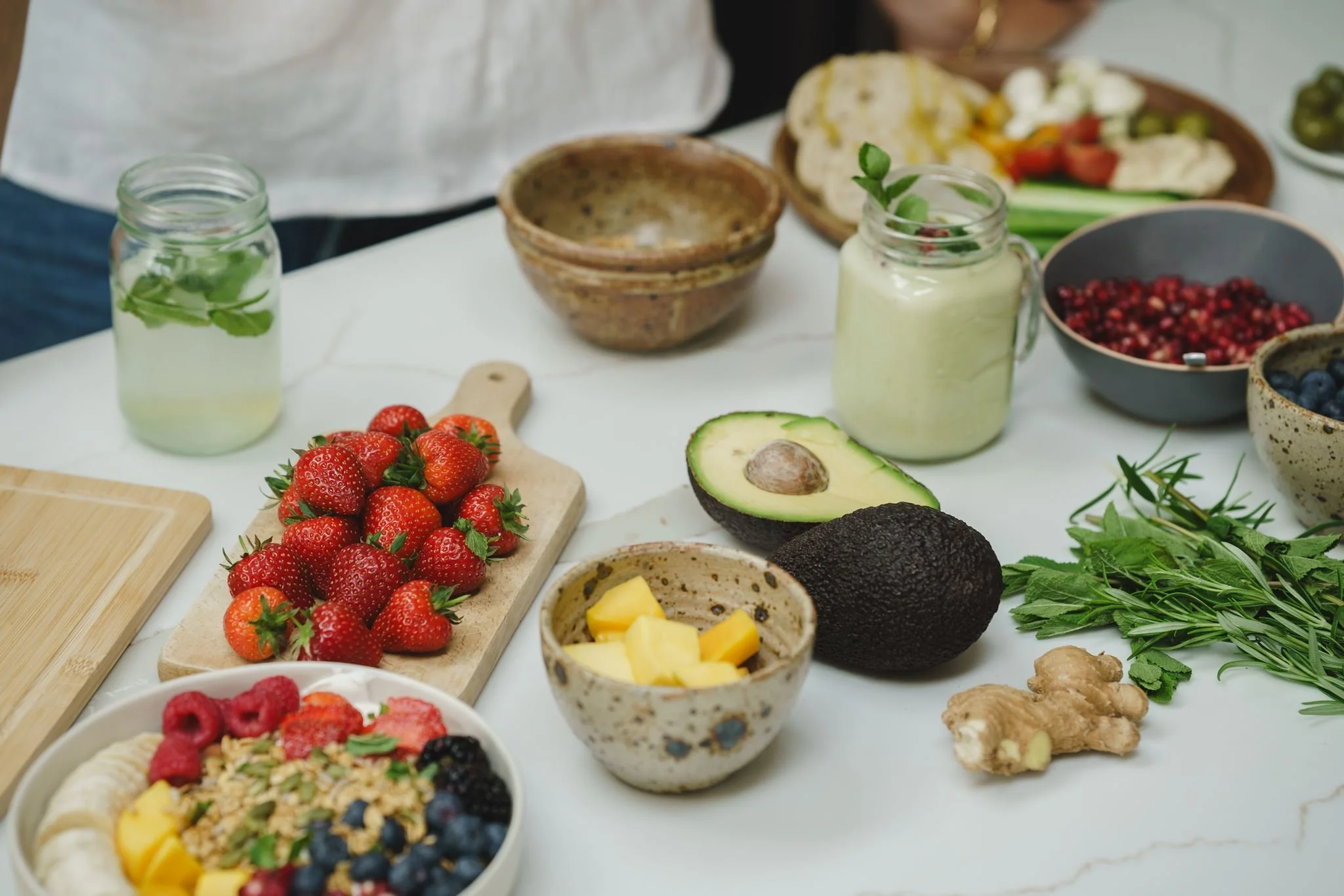 A spread of fresh fruits, herbs, smoothies, and snacks on a white table, including strawberries, avocado, blueberries, pomegranate, ginger, and a smoothie in a glass jar.