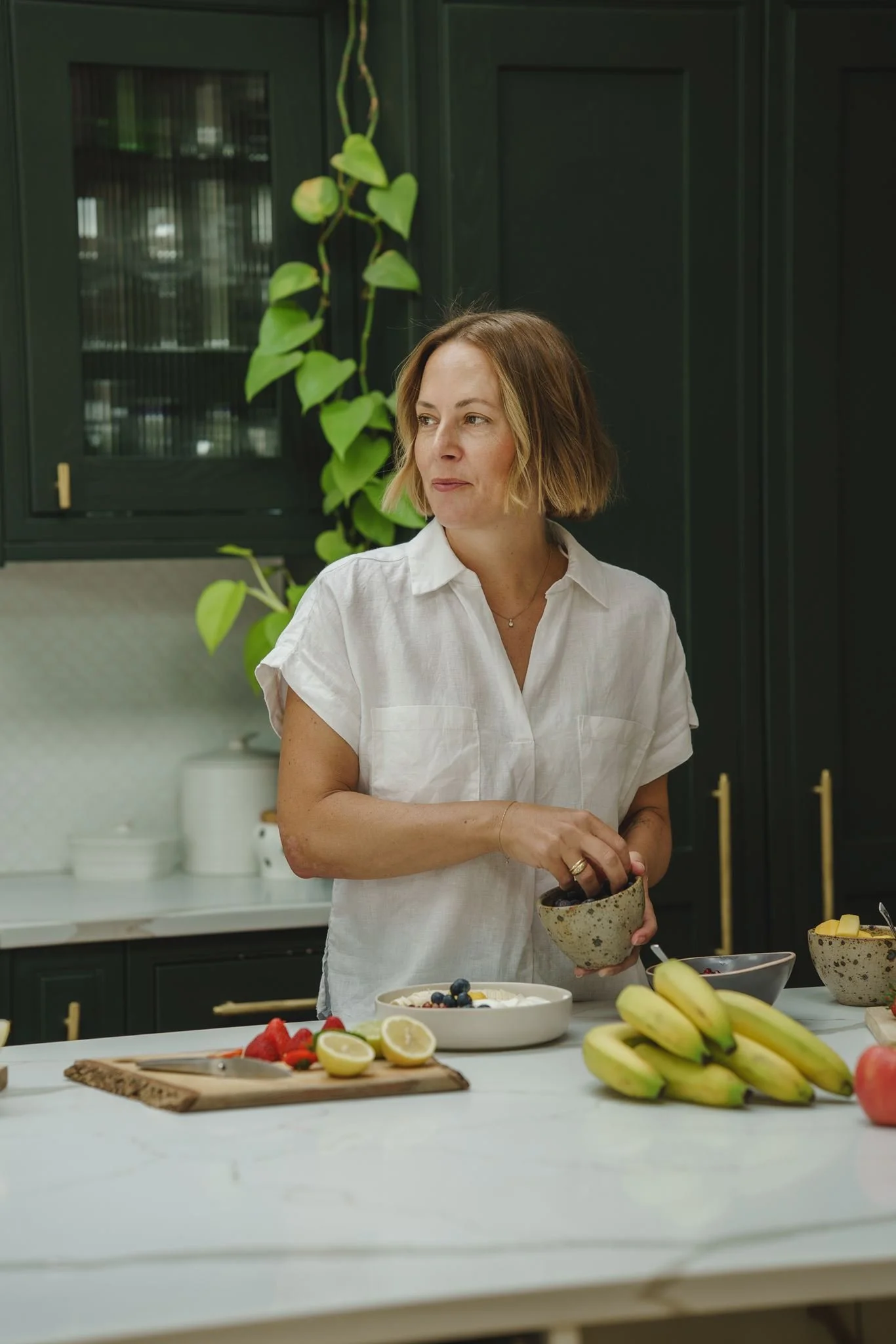 A woman with shoulder-length blonde hair in a white shirt is standing in a kitchen, holding a ceramic bowl. The kitchen has dark green cabinets, a white countertop, and a window with vertical blinds. There are various bowls of fruit and a wooden cutting board with sliced fruit on the counter.
