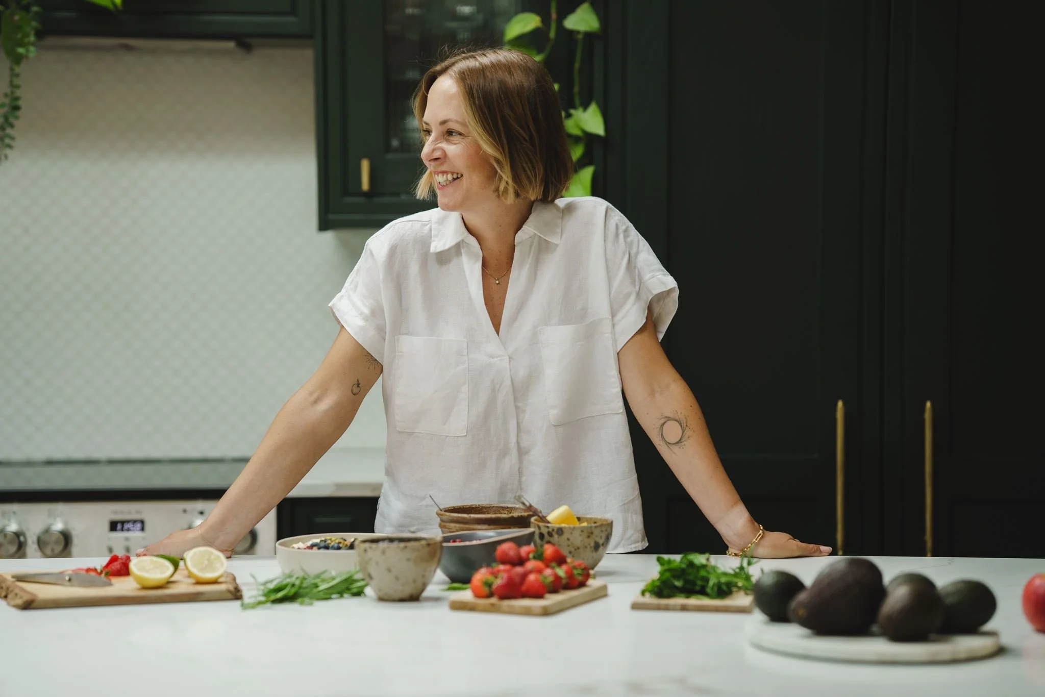Woman smiling at a kitchen counter with bowls of strawberries, avocados, and lemons, and green herbs and vegetables scattered on the counter.