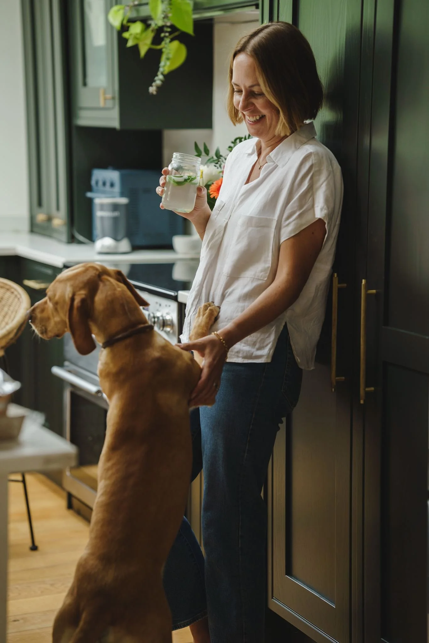 A woman with shoulder-length brown hair, wearing a white shirt and jeans, is smiling and holding a jar of water decorated with lime slices and mint leaves. She is standing in a kitchen next to a brown dog that is sitting and looking up at her, with its front paws resting on her lap.