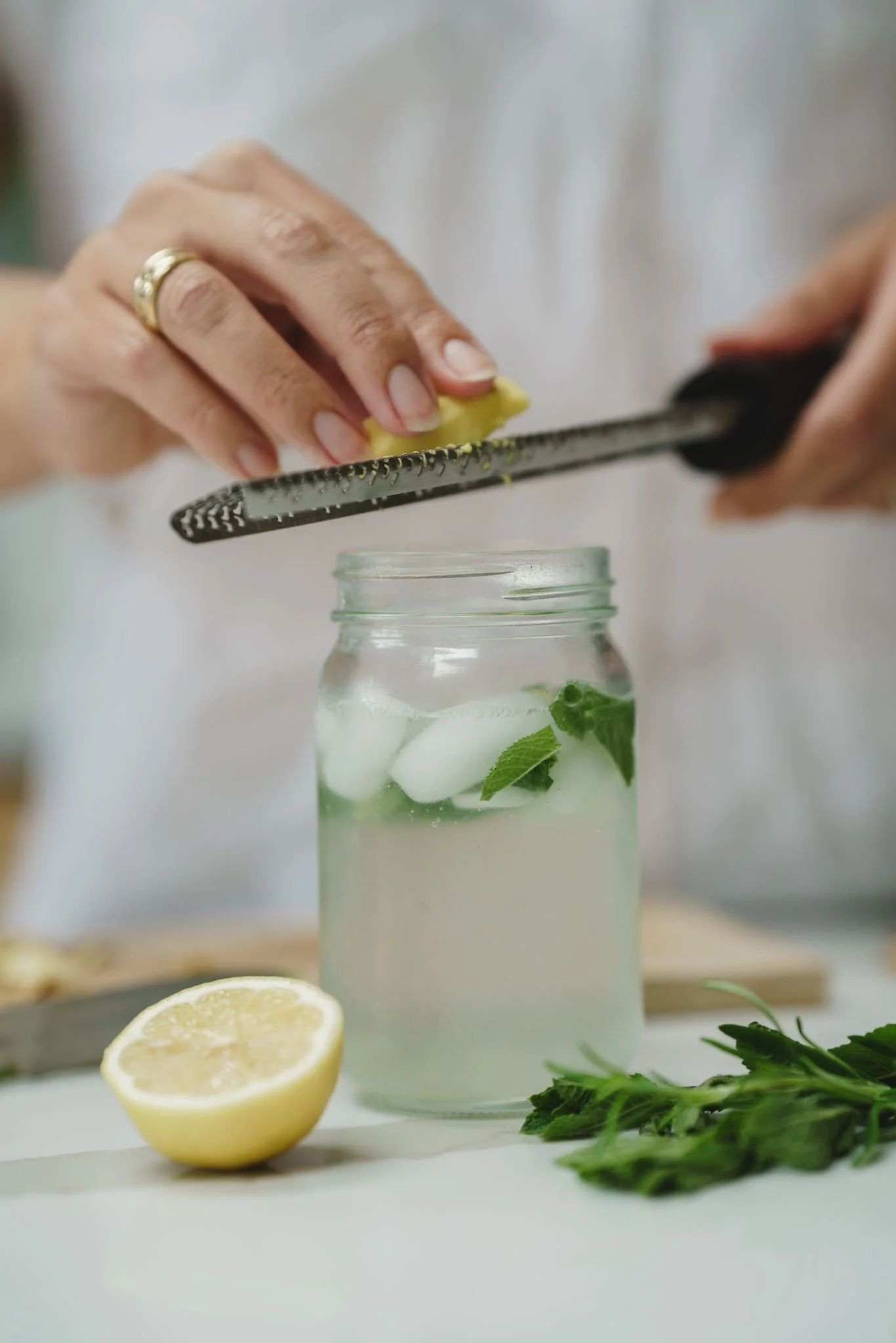 Person grating a lemon above a jar of water with mint leaves and ice, with lemon halves and fresh mint on the table.