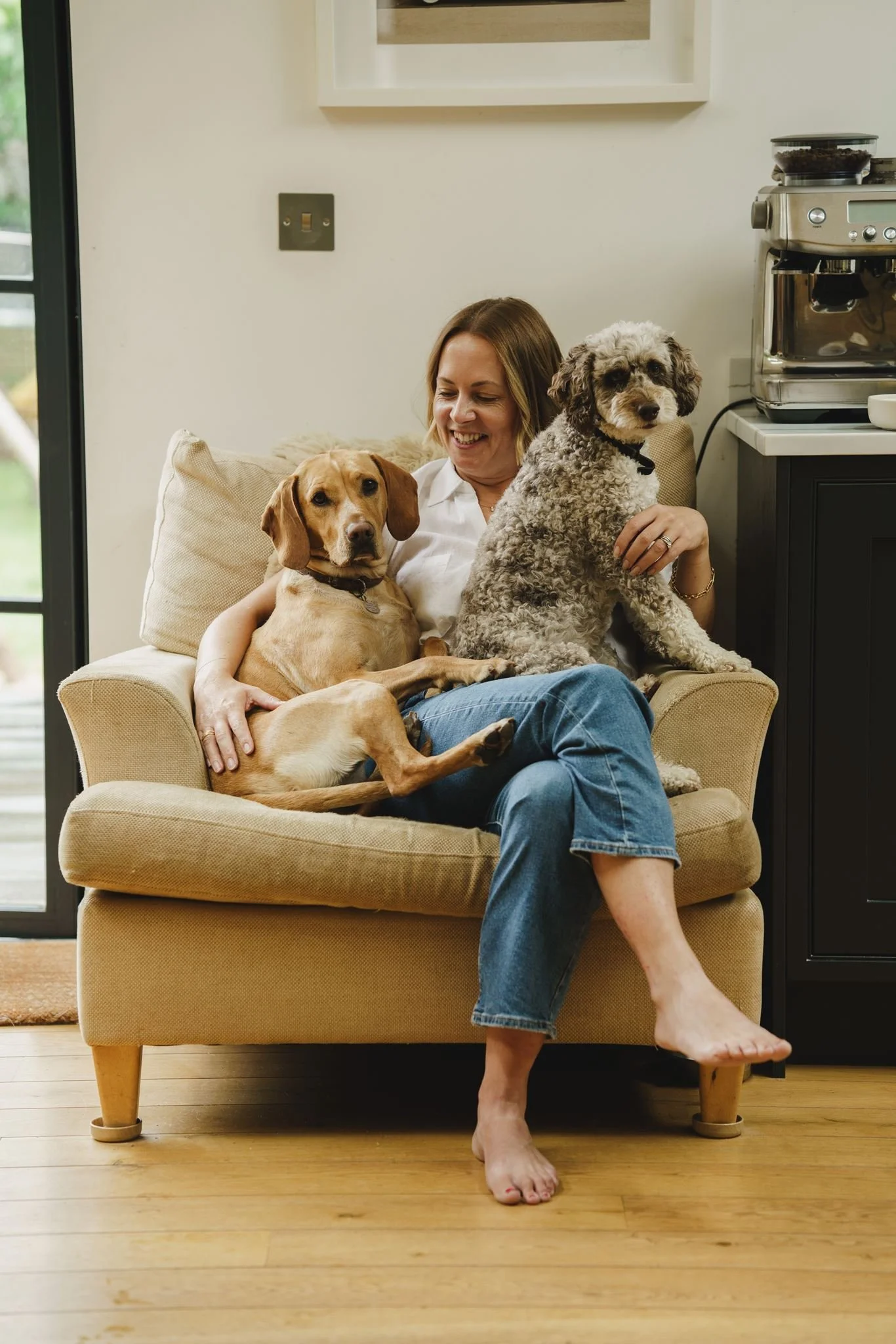 A woman sitting on a beige armchair with her legs crossed, holding and hugging two dogs, one sitting on her lap and the other on her shoulder. She is smiling and looking down at the dog on her lap. The room has a wooden floor, a small dark cabinet, a coffee machine, and a window with a view outside.