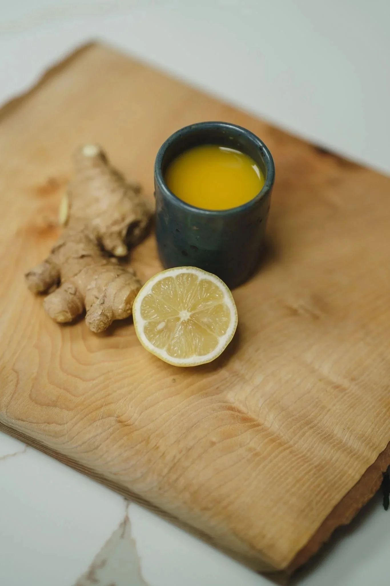 A wooden cutting board with a sliced lemon, a piece of ginger root, and a small black cup filled with yellow liquid.