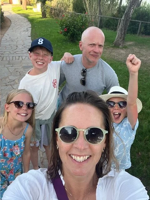 Family of five outdoors, smiling for a selfie, with a woman in the center wearing sunglasses, a man behind her, two girls wearing sunglasses and hats, and a boy wearing a baseball cap.