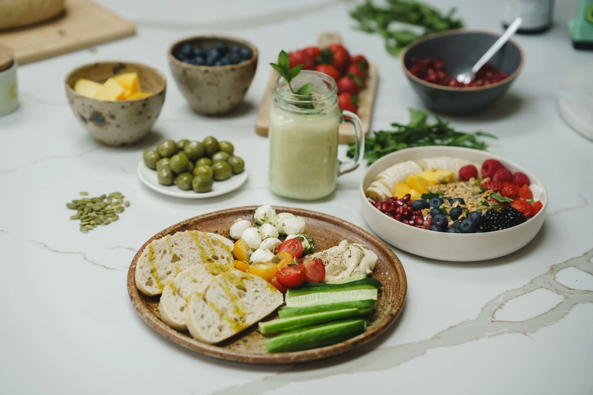 Assorted fresh fruits, vegetables, and a smoothie on a white marble countertop, including a bowl of blueberries, strawberries, raspberries, blackberries, green olives, sliced bread, cucumber sticks, and a bowl of granola with berries.