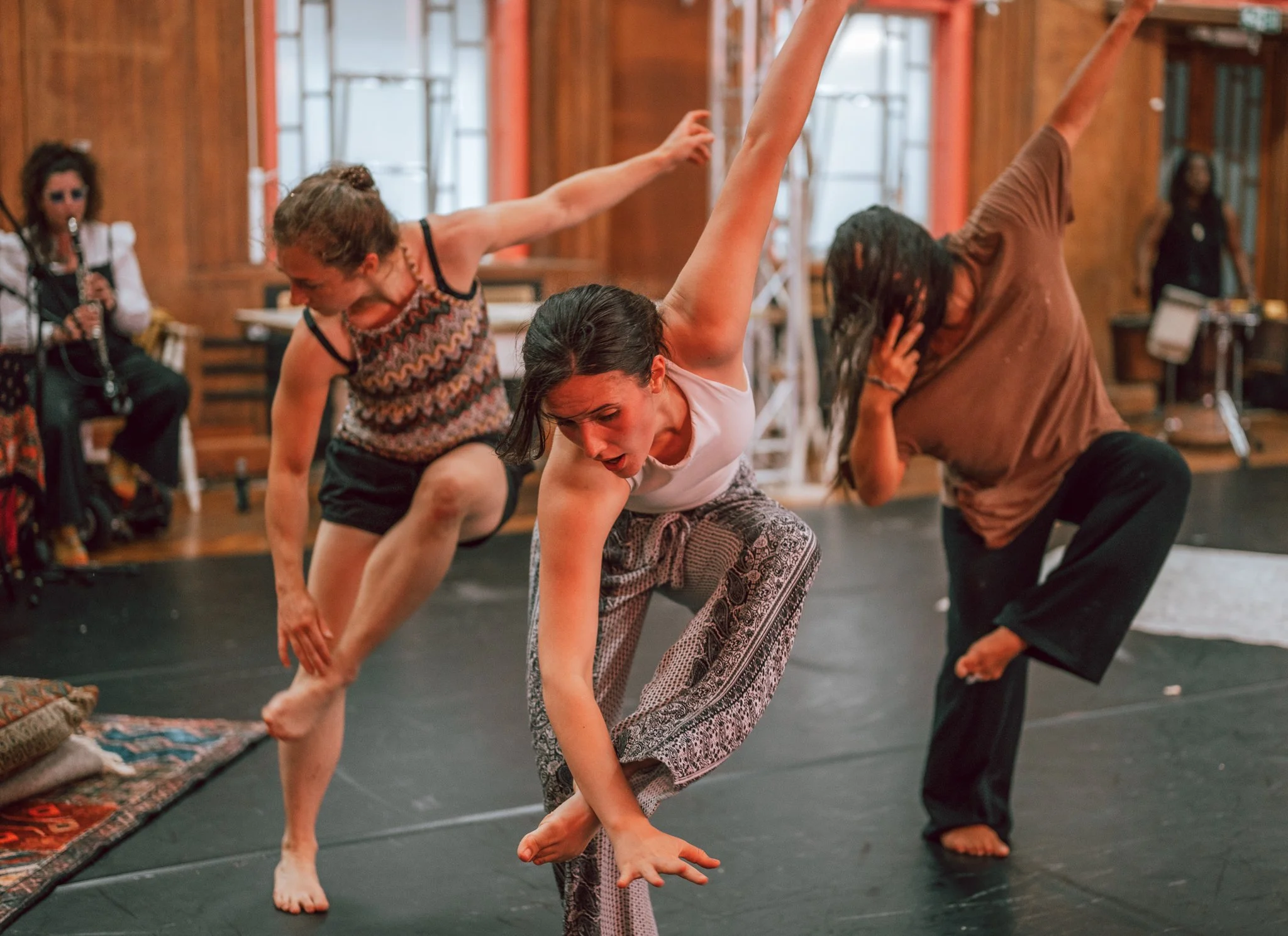 Two women are practicing acro yoga or partner balancing exercises indoors, with one woman supporting the other as she balances on her hands and feet. Other people are visible in the background, watching or waiting.