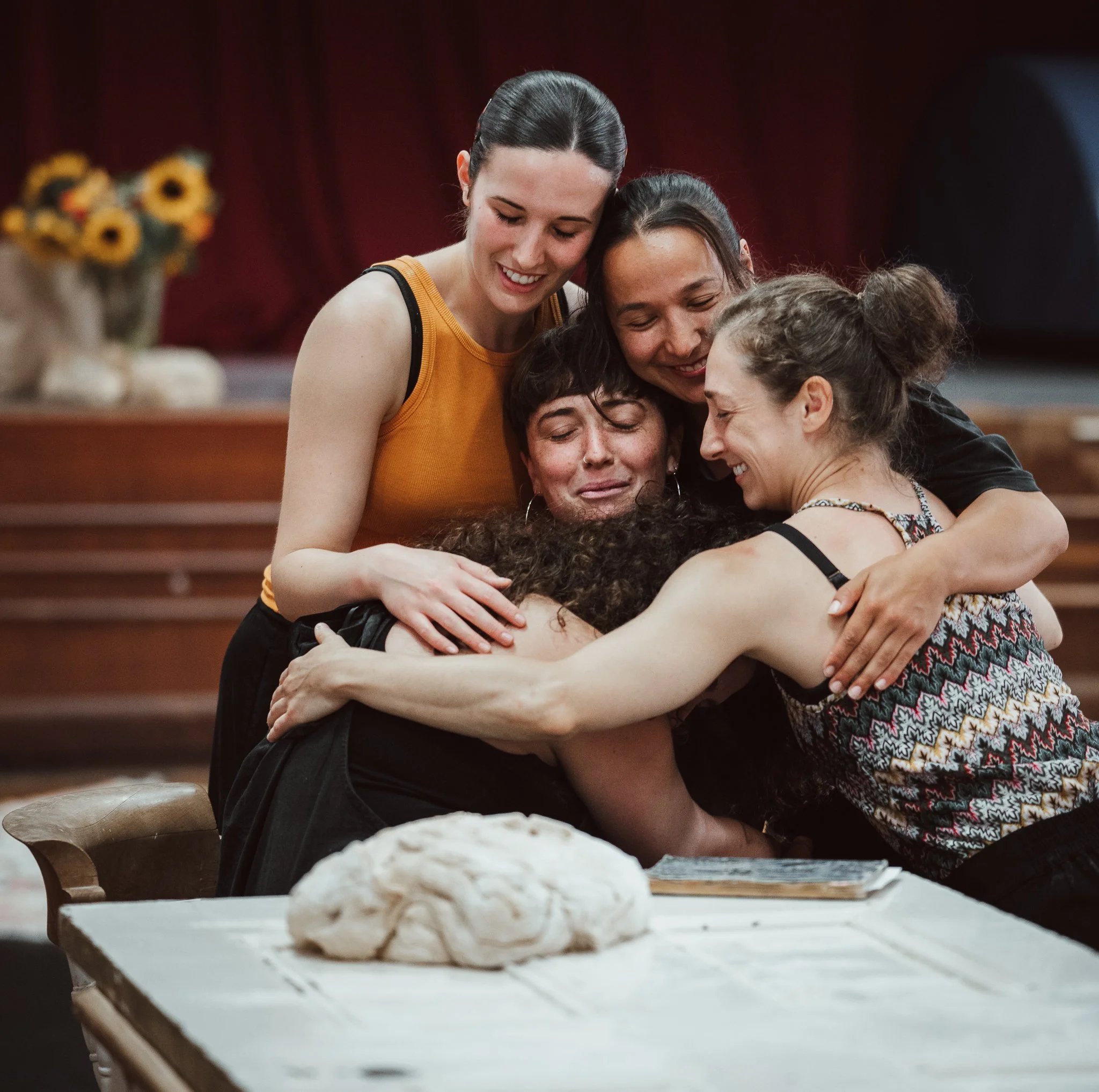 Seeking dance performers hug each other in an indoor setting, with a table of bread in the foreground and a sunflower decoration in the background.