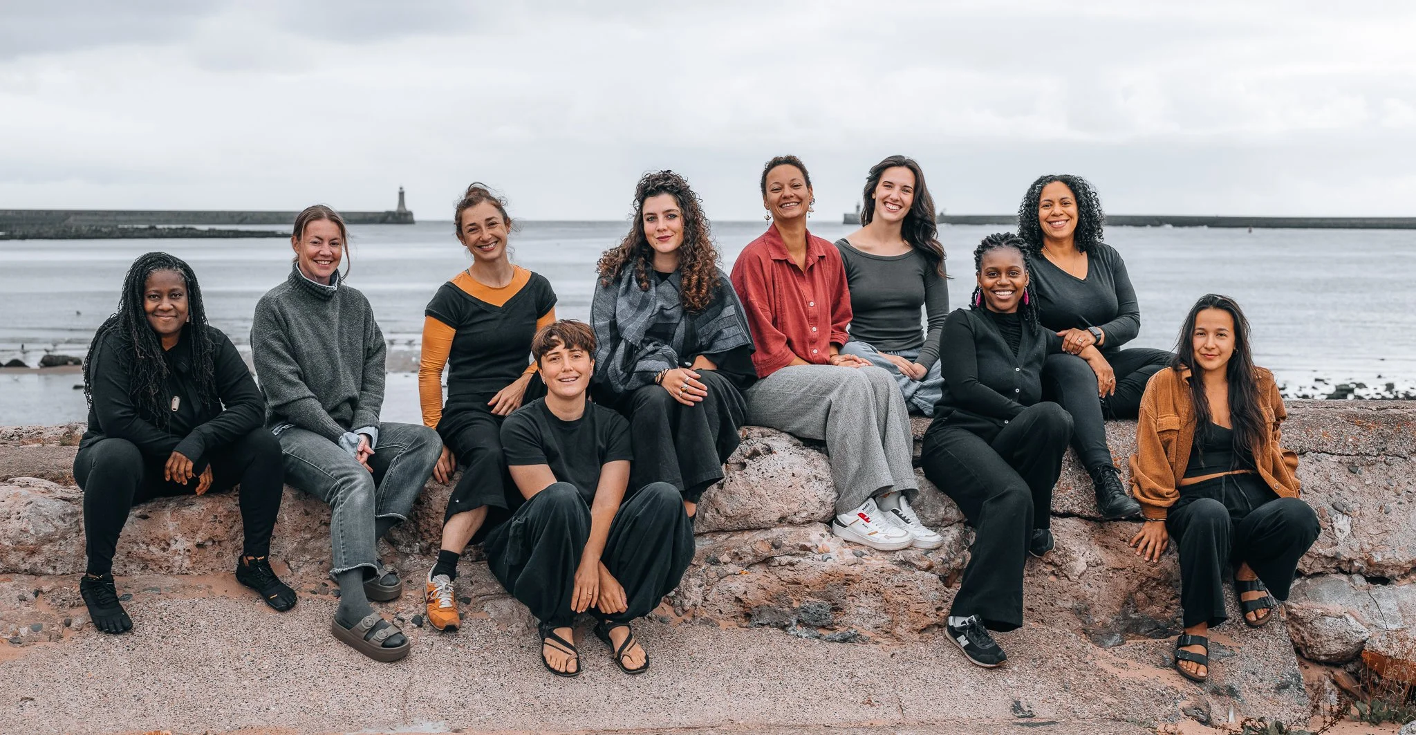 Group of diverse women sitting on rocks by the ocean with lighthouses in the background amid a cloudy sky.
