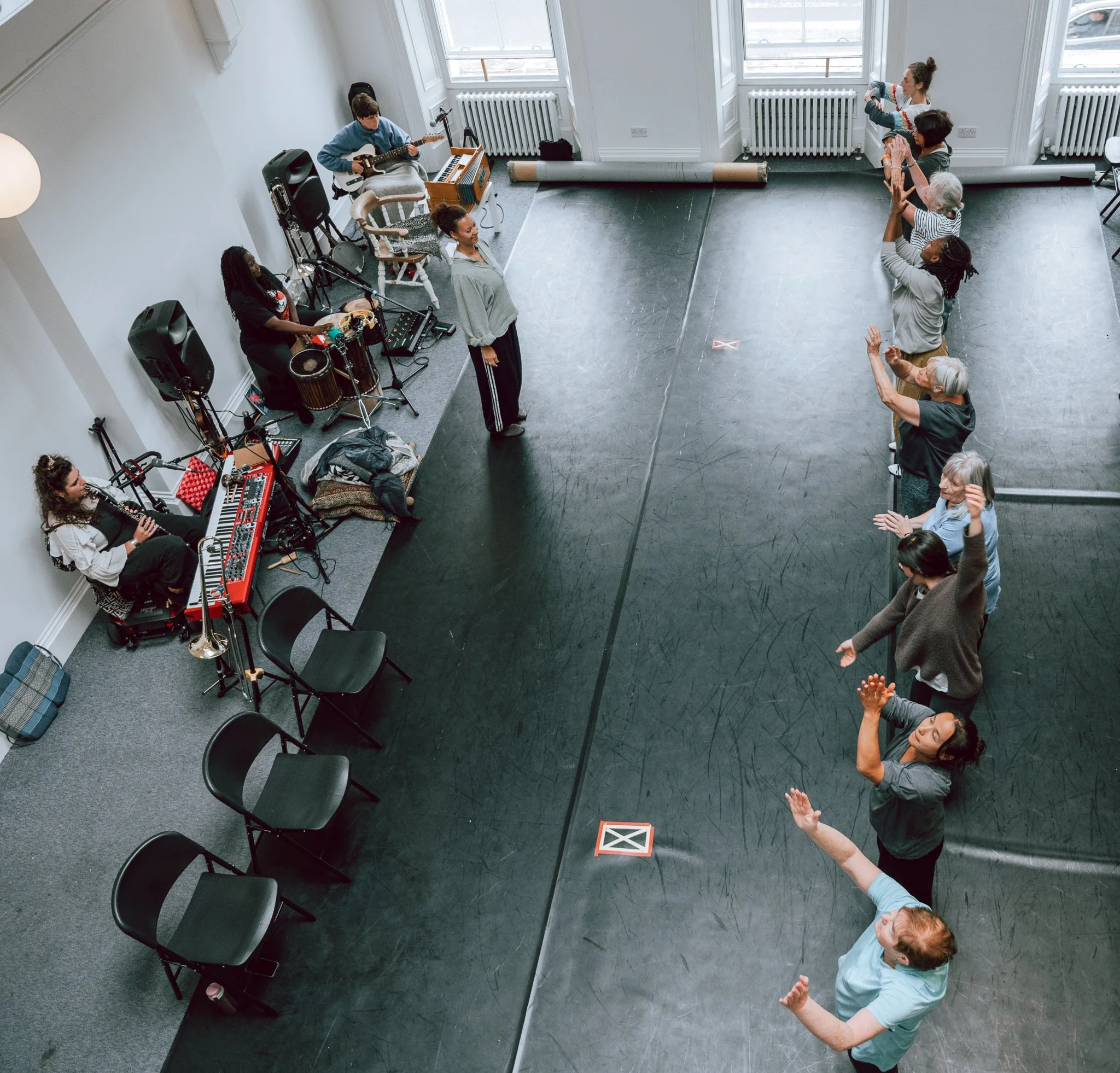 A group of people participating in a dance class in a spacious, well-lit room with large windows. Some are standing and following the instructor, while others are sitting and playing musical instruments including a keyboard, a drum, and a guitar.