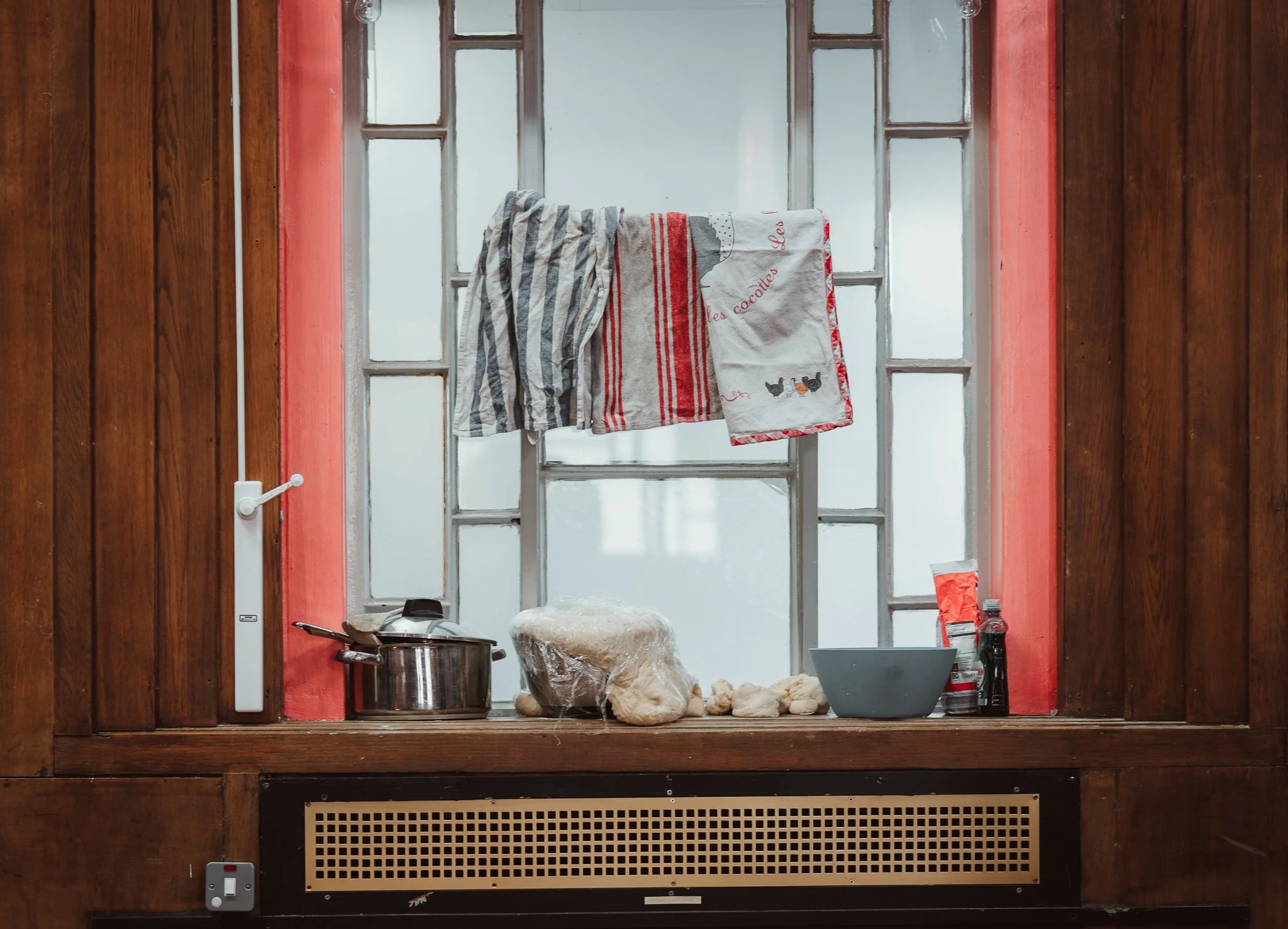 Kitchen window with wooden frame, hanging dish towels, cooking pot, food items, and kitchen utensils on the windowsill.