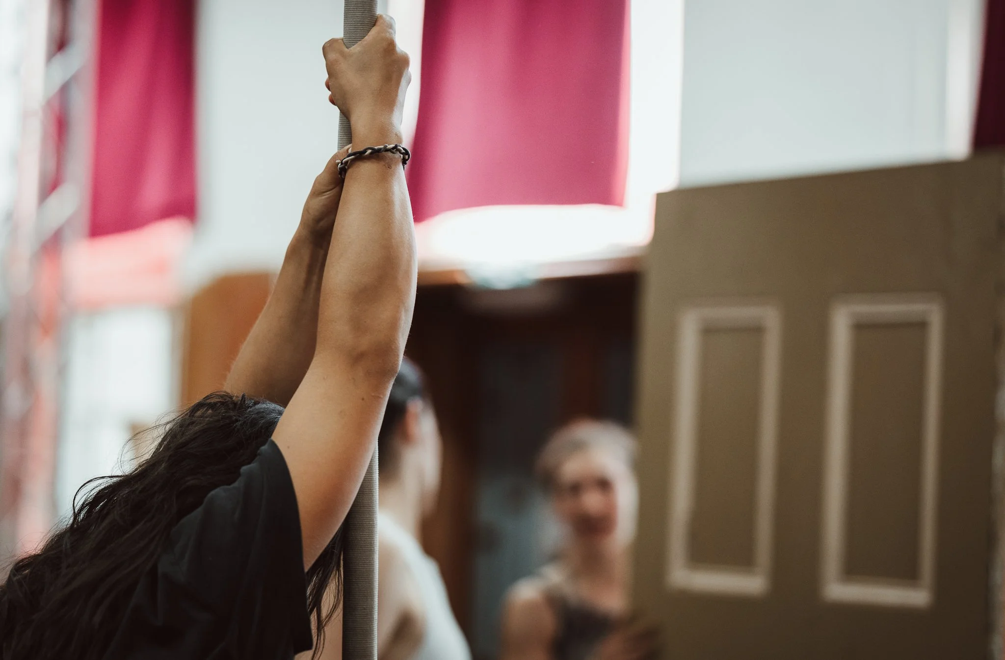 A person with long dark hair wearing a black short-sleeved shirt is raising both hands, gripping a ballet barre in a dance studio. Other people and windows are visible in the background.