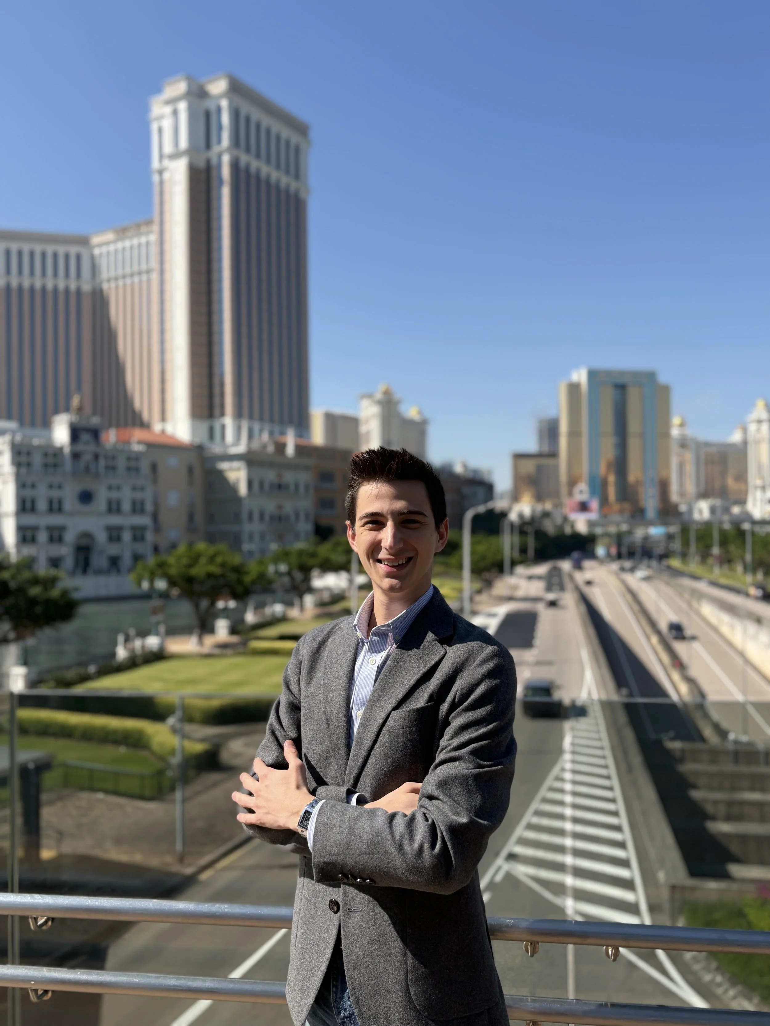 Young man in gray blazer smiling with arms crossed on a balcony overlooking city streets and tall buildings on a sunny day.