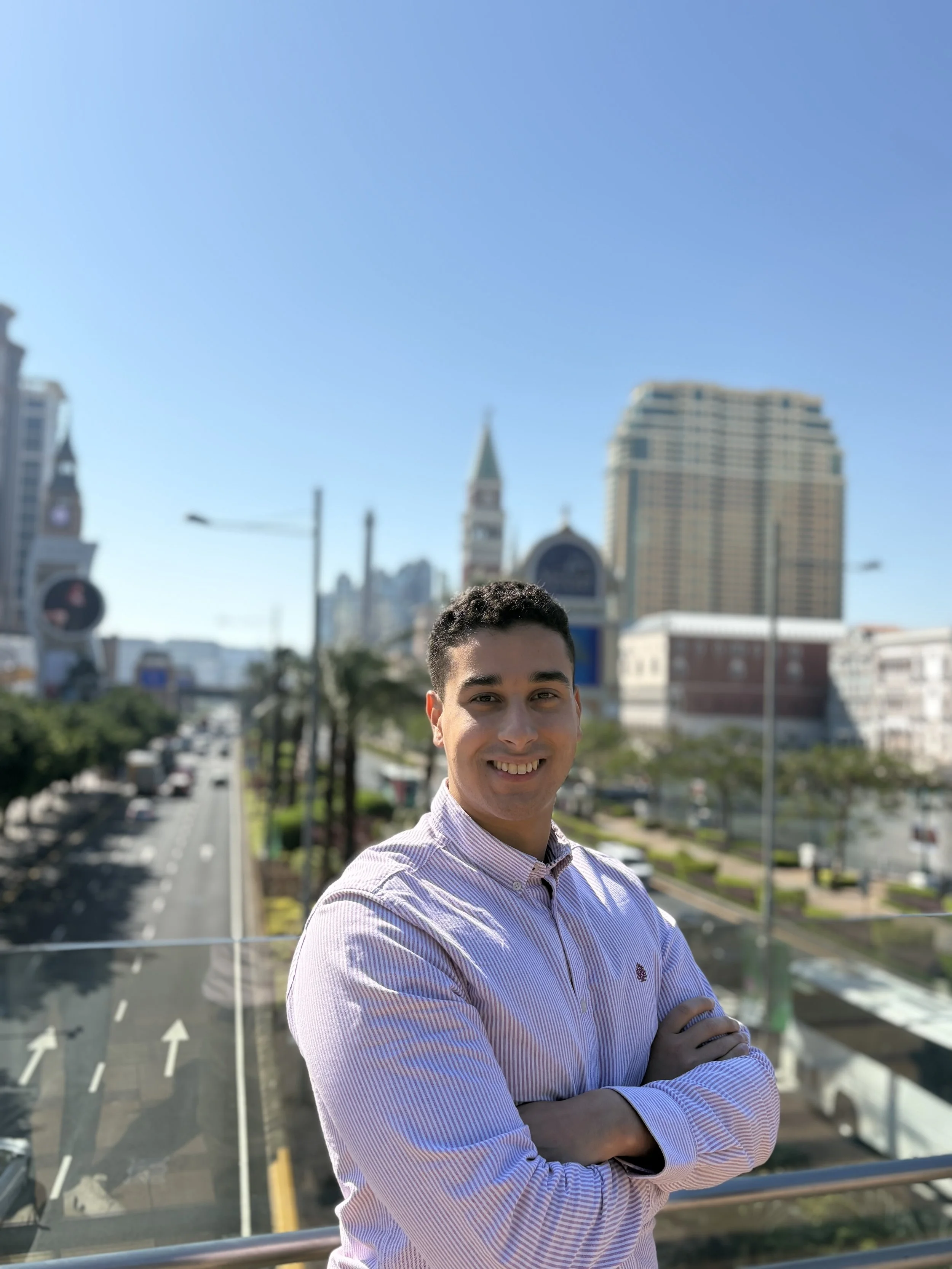 Smiling young man with arms crossed on a balcony in an urban city with tall buildings and a clock tower in the background, under a clear blue sky.