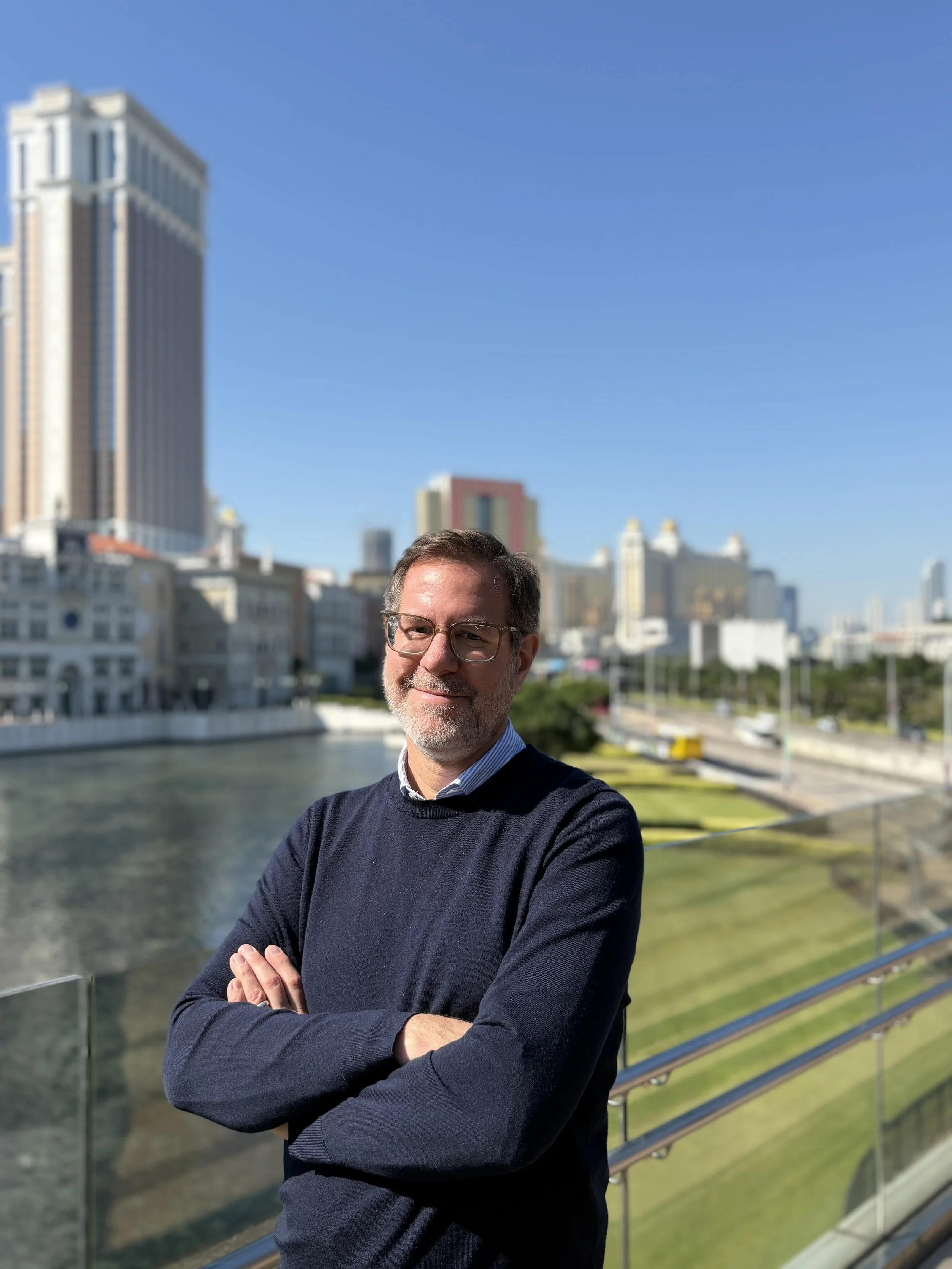 A smiling man with glasses and a gray beard, wearing a navy sweater over a collared shirt, standing on a balcony with crossed arms, overlooking a city skyline with tall buildings, a river, and a green park, on a sunny day.