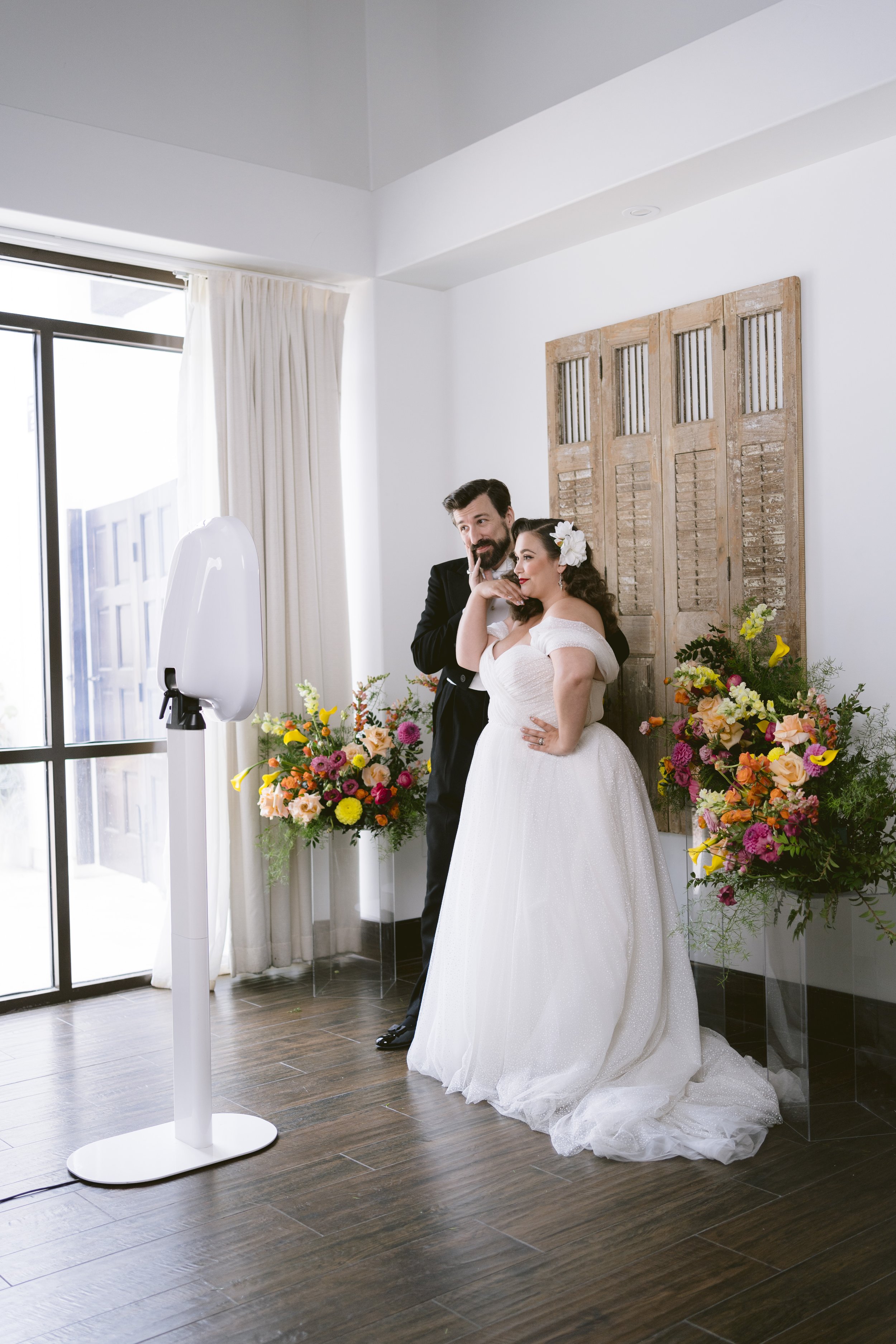 A bride and groom dressed in wedding attire stand together in a bright room with large windows, colorful flower arrangements, and a wooden decorative window shutter on the wall.