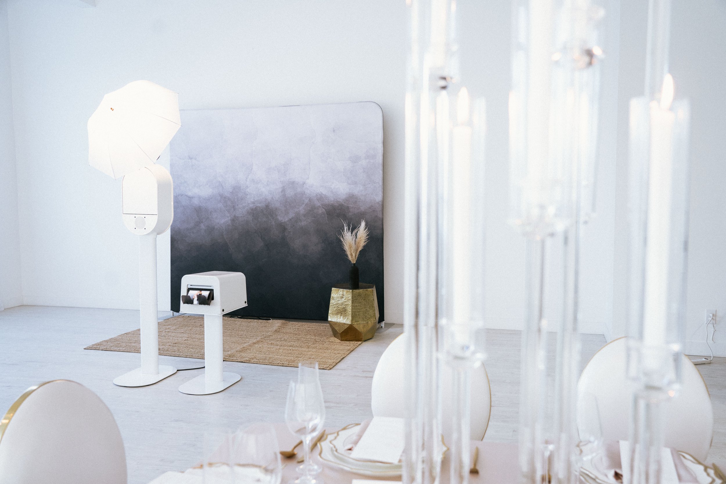 Elegant banquet or event space with a white table set with glassware and gold accents, blurry in the foreground, with modern decor including a black and white backdrop, a tall white lamp, a small white machine, and a gold geometric vase with pampas grass on a woven rug against a white wall.