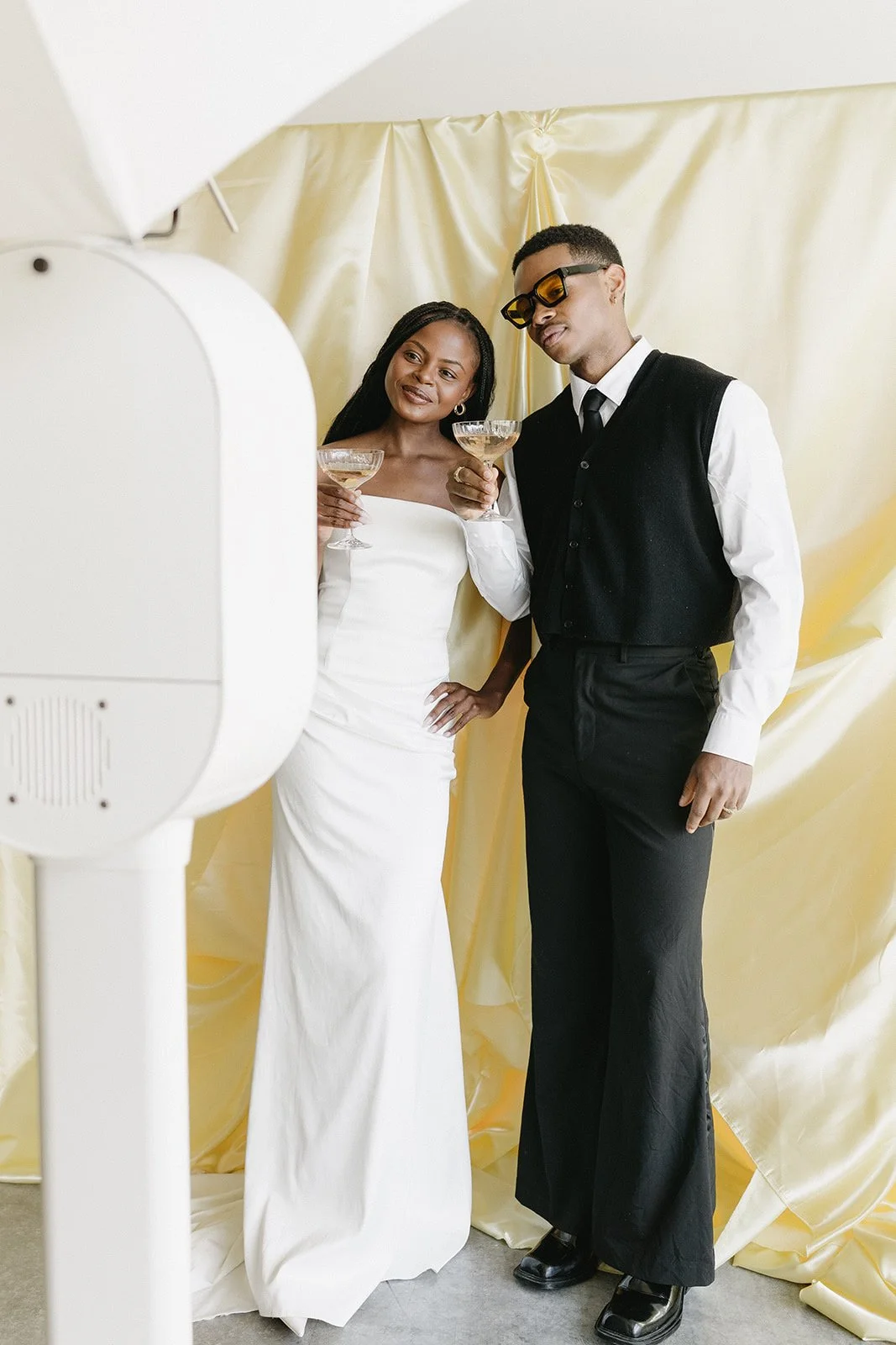 A woman and man take a photo together, both holding champagne glasses. The woman is wearing a white dress, and the man is dressed in a black vest over a white shirt, with sunglasses.