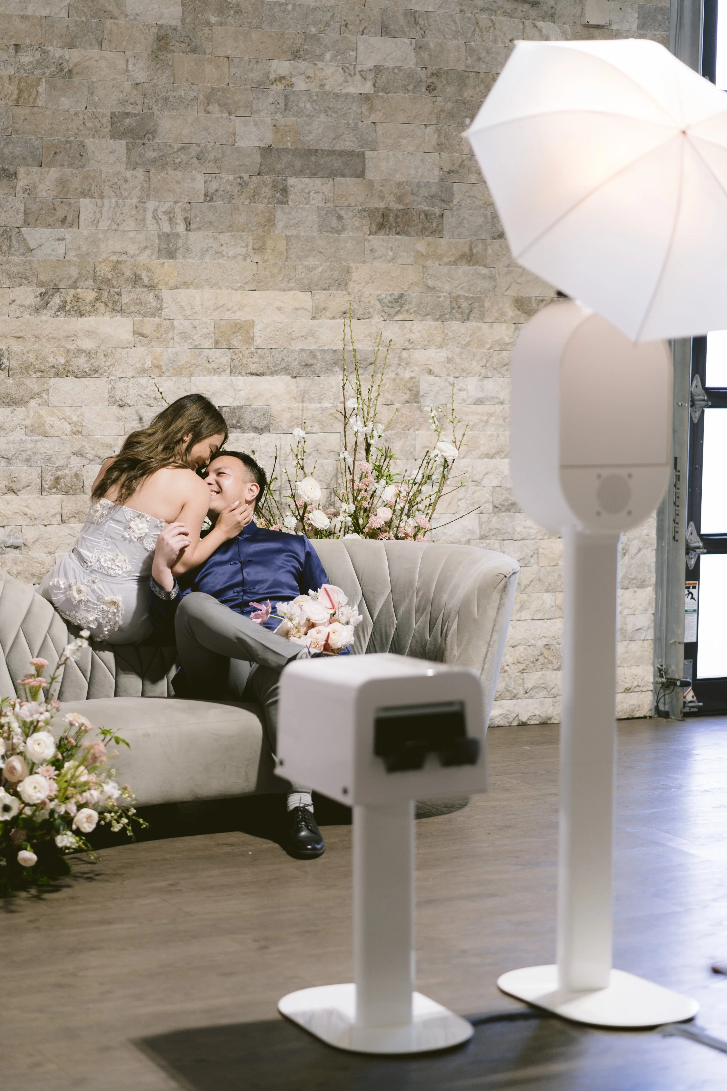 A couple is seated on a gray sofa, with the woman leaning over the man, both smiling and looking happy, surrounded by flower arrangements and a stone wall background. In the foreground, there are two white, modern medical or dental devices.
