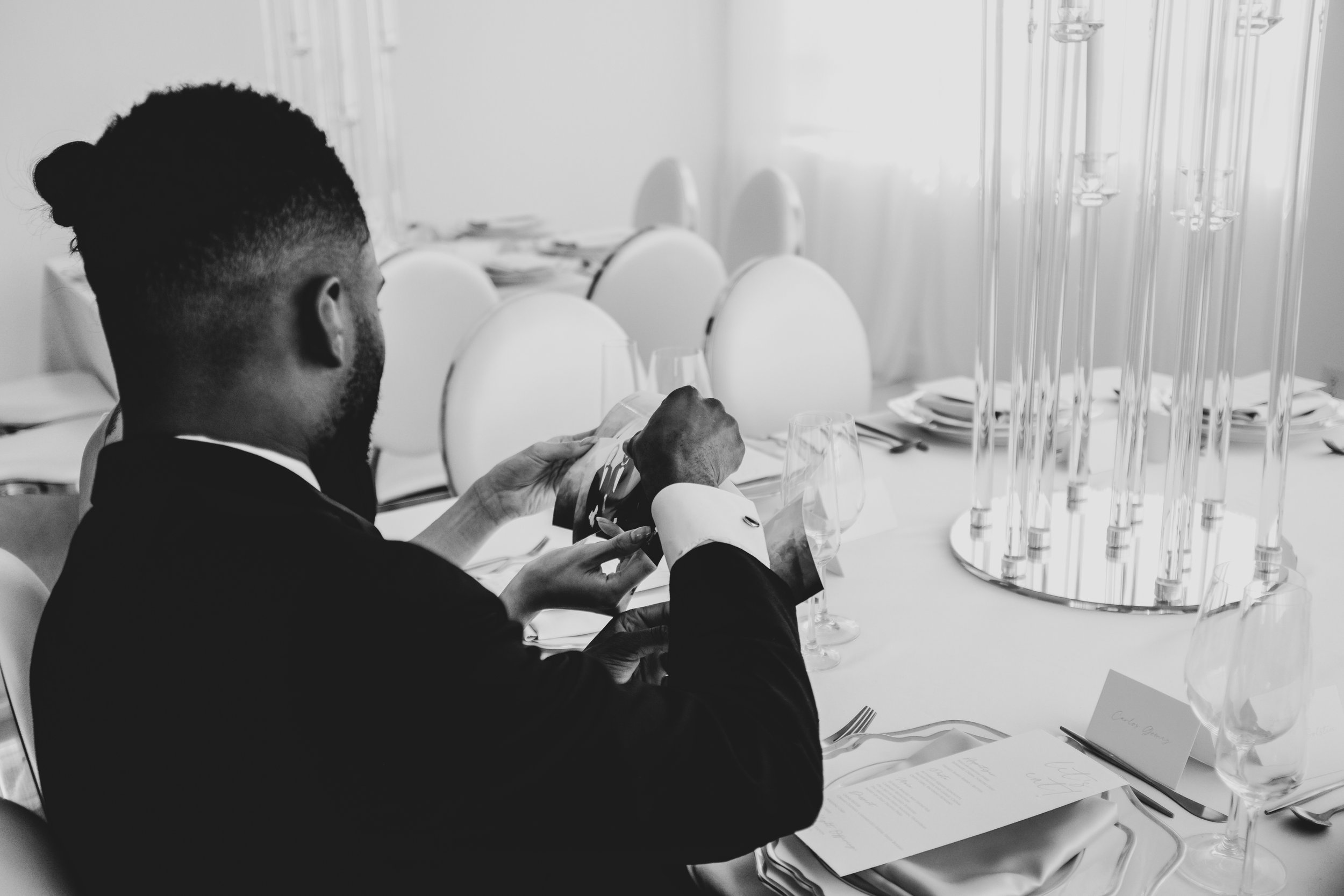 Male in black and white dressed in formal clothes holding a photobooth photo discussing with guest at dinning table
