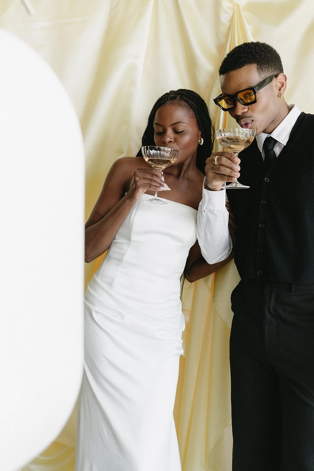 A woman in a white dress and a man in a black vest and glasses drinking champagne together at an event.