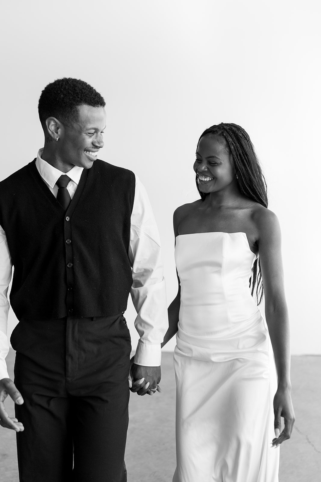 A black-and-white photo of a smiling couple holding hands and walking together. The man is wearing a white shirt with a black vest and black pants, and the woman is dressed in a strapless white dress.