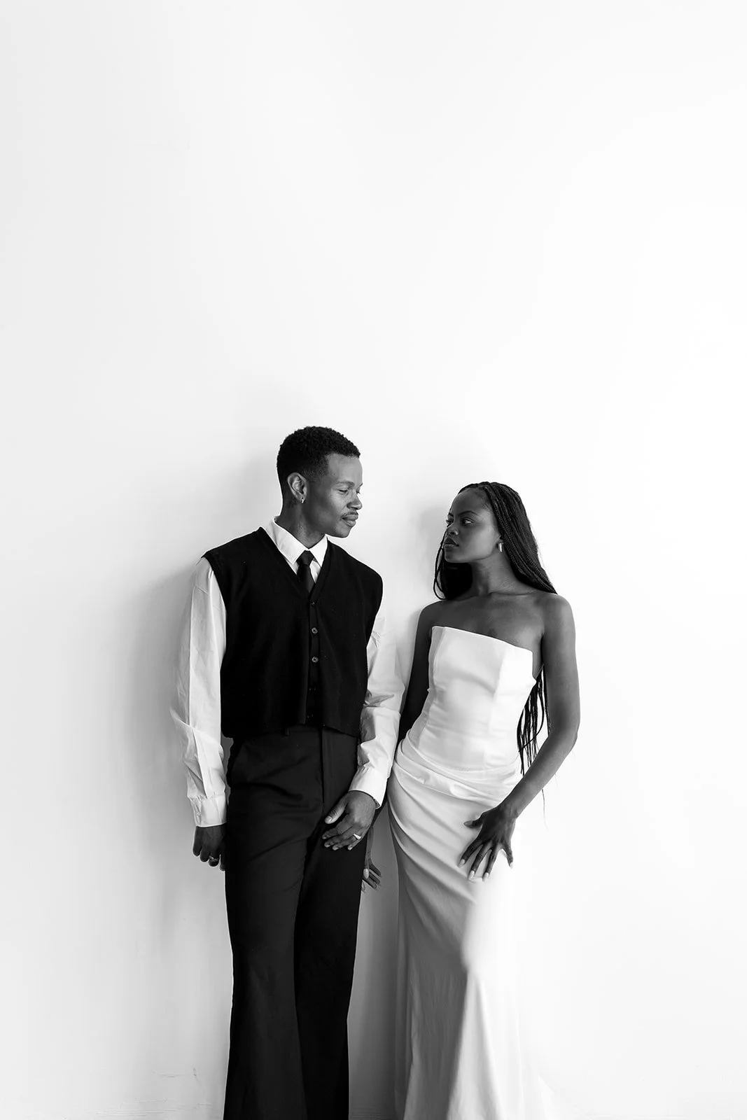 Black and white photo of a man and woman standing against a plain white wall, looking at each other. The man is wearing a white shirt, black vest, and black pants. The woman is wearing a strapless, fitted white dress with long braids.