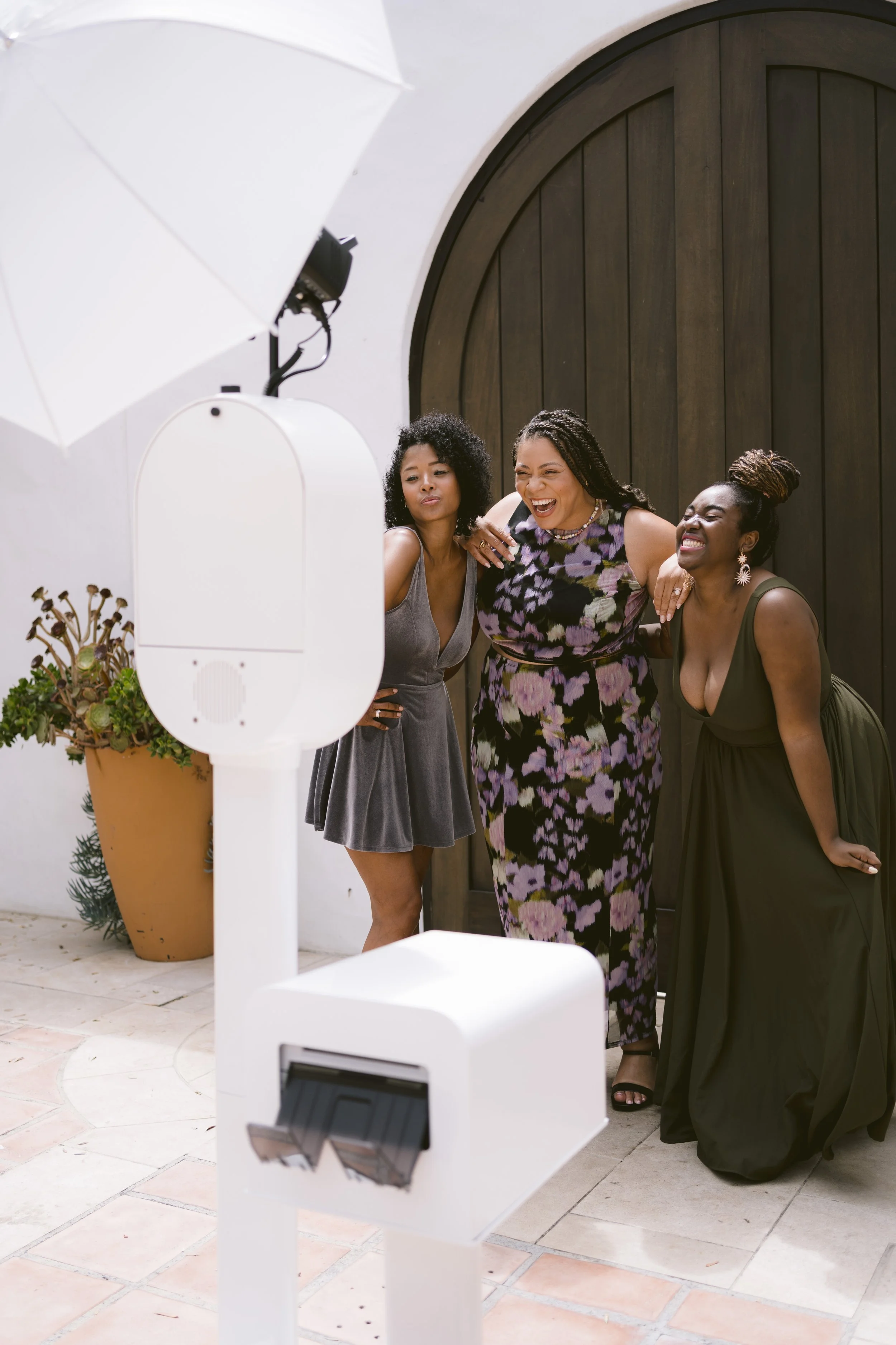 Three women laughing and posing for a photo outside near a large dark wooden door, with photography equipment including a softbox and a plant in a large terracotta pot visible in the foreground.