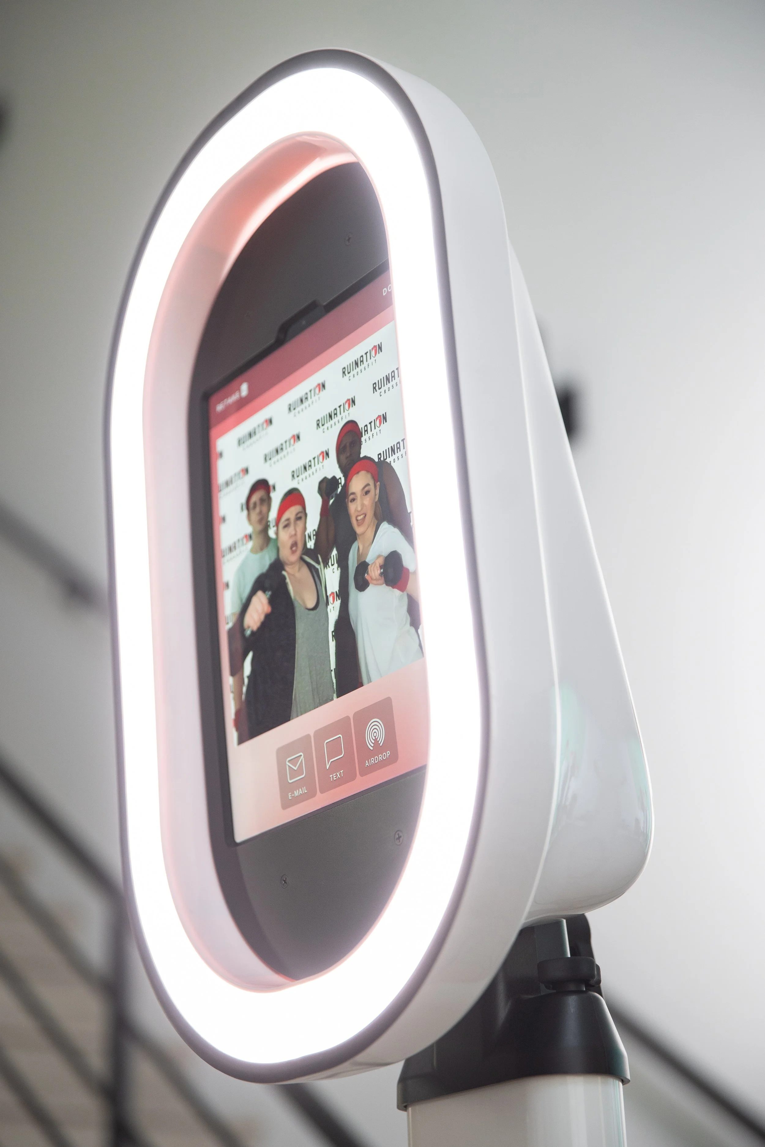 Photo of a modern fitness kiosk with a touchscreen display showing four people exercising with dumbbells, a digital interface, and icons for email, text, and AirDrop, surrounded by a circular lighting ring.