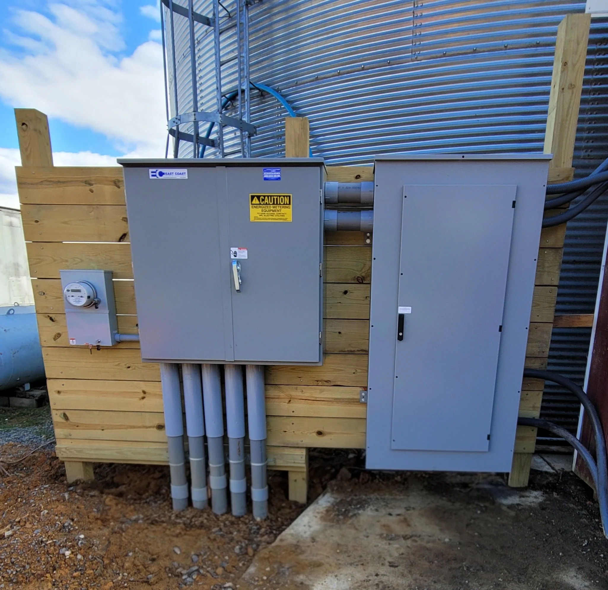Electrical panels mounted on a wooden platform outside, with conduit pipes and a utility meter visible.