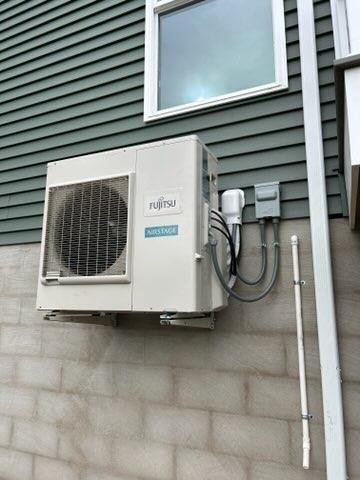 Outdoor Fujitsu air conditioning or heat pump unit attached to the exterior wall of a building, with electrical wiring and pipes connected.