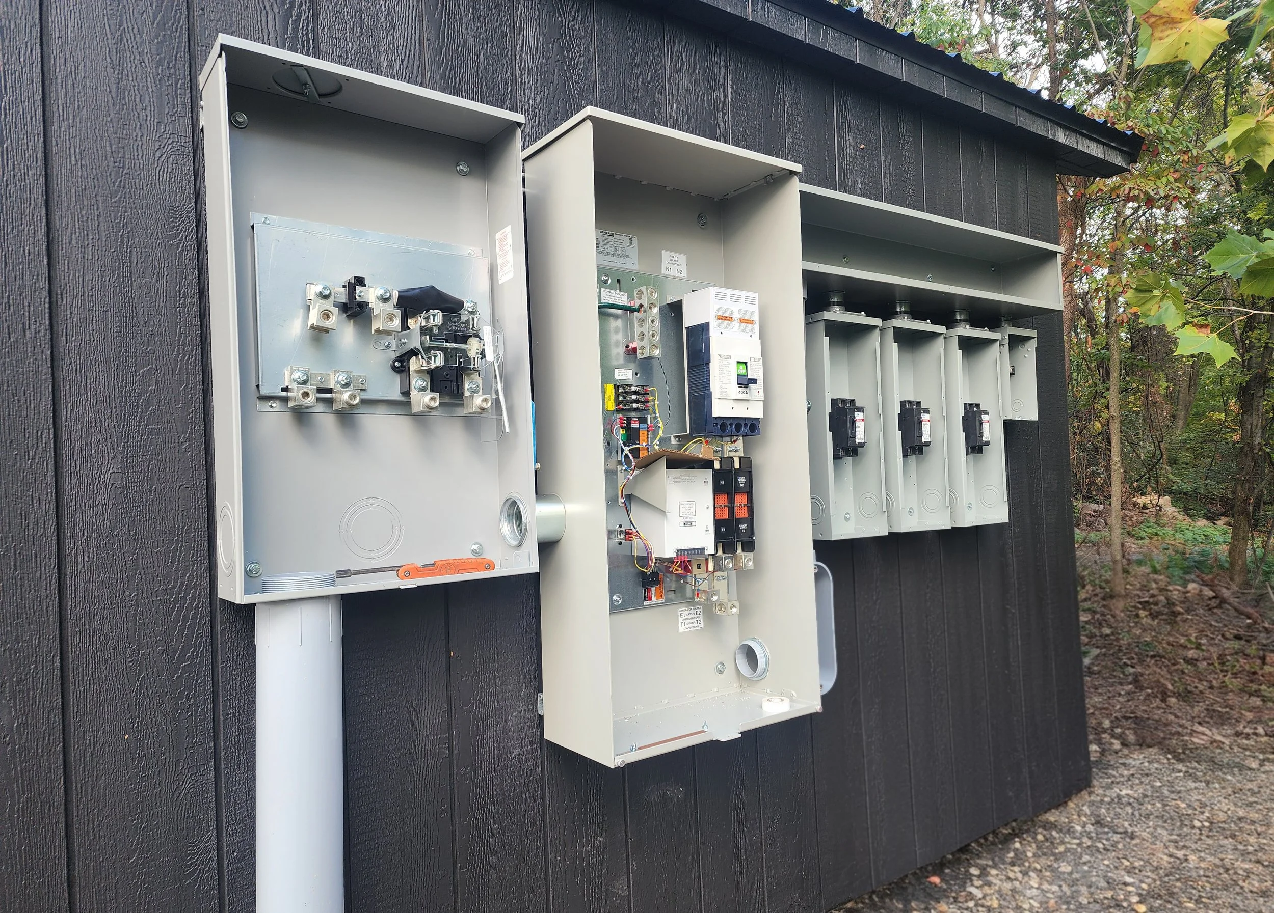 Outdoor electrical panel boxes mounted on a dark wooden wall, with some panels open showing internal wiring and circuit breakers, surrounded by trees and leaves.