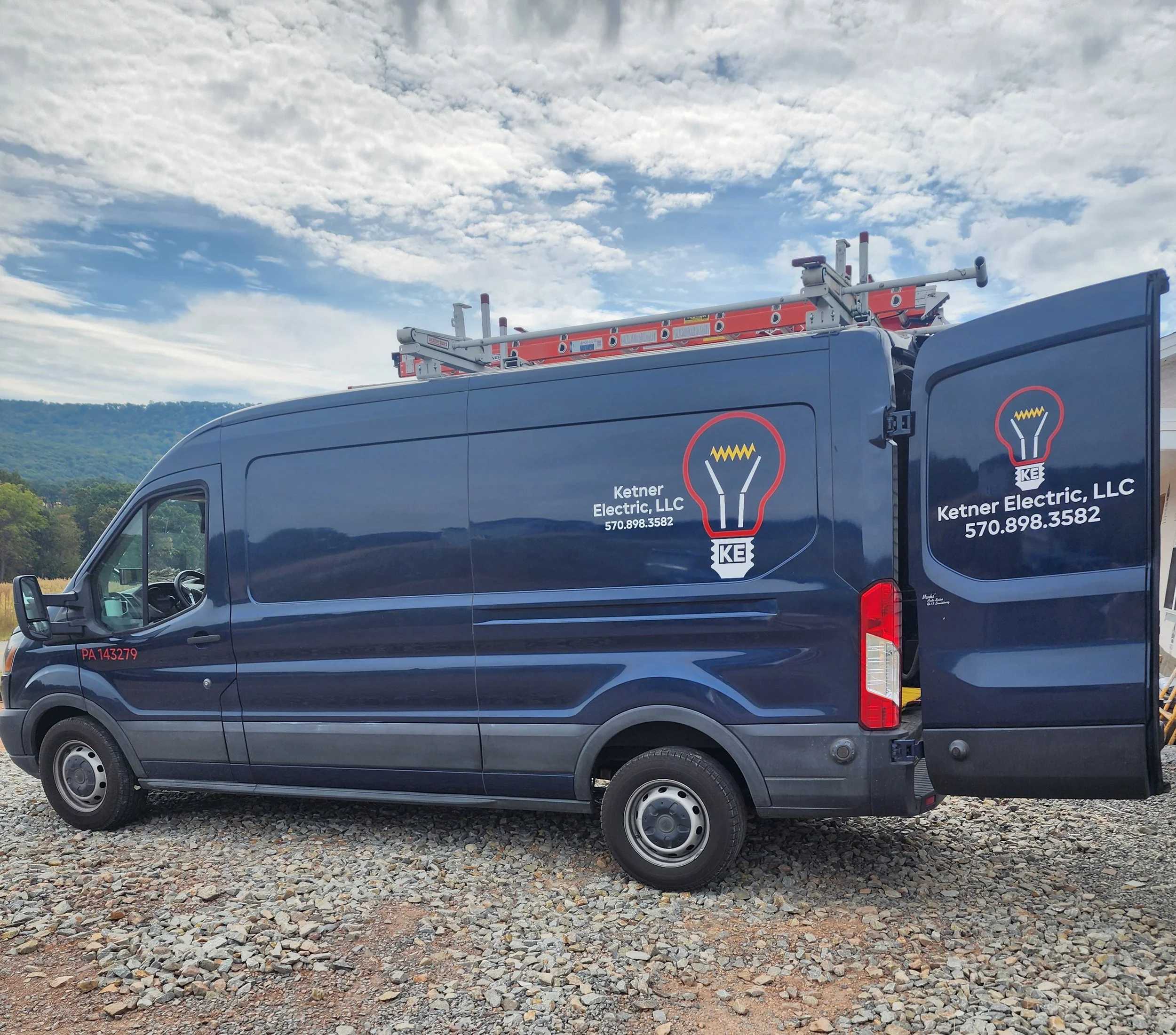 A blue company van with the logo and contact information of Ketner Electric, LLC, parked on a gravel surface with a cloudy sky and hills in the background. The van has a ladder on its roof and the right rear door is open.