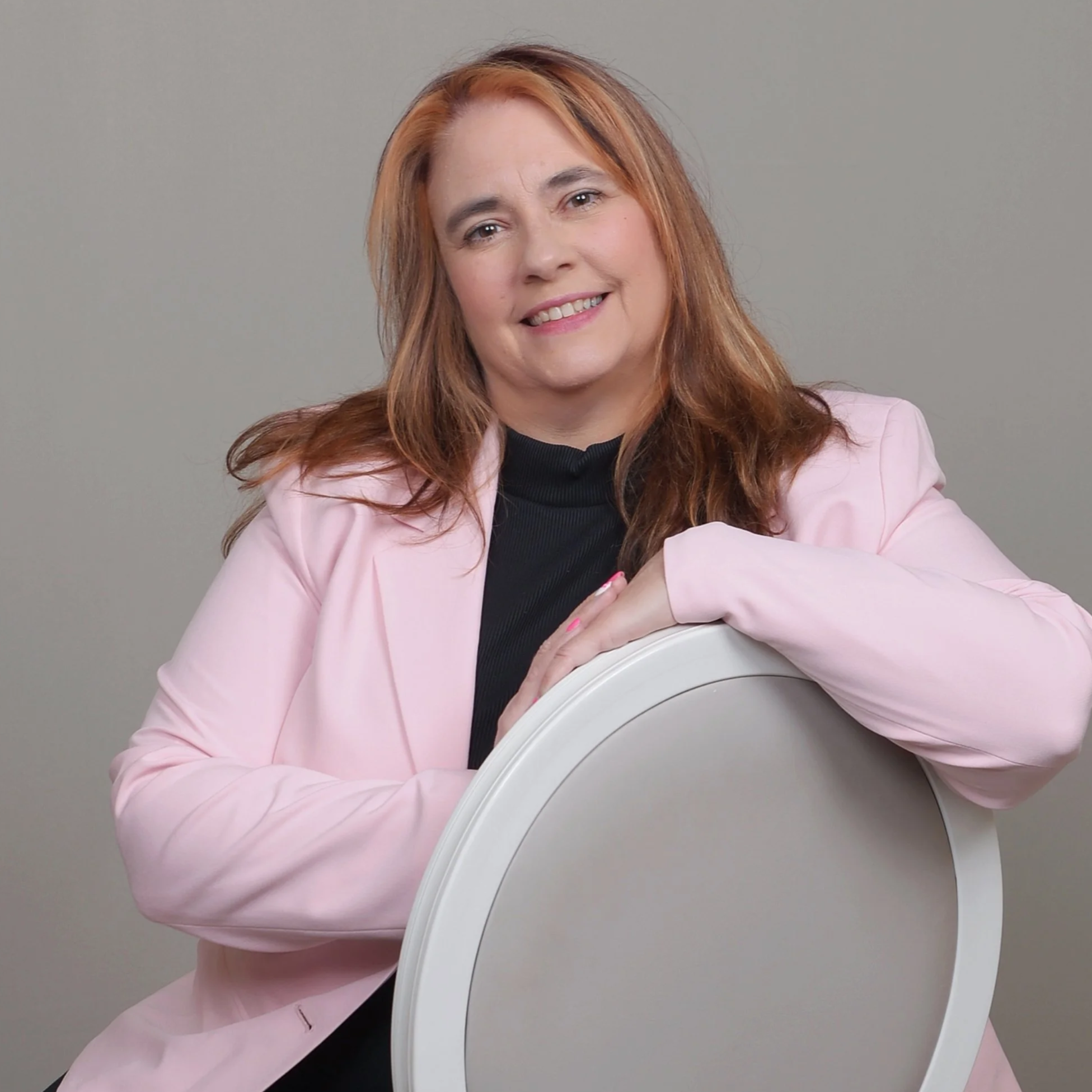 A woman with red hair wearing a pink blazer and black top, smiling and resting her arm on a white chair, against a plain light gray background.