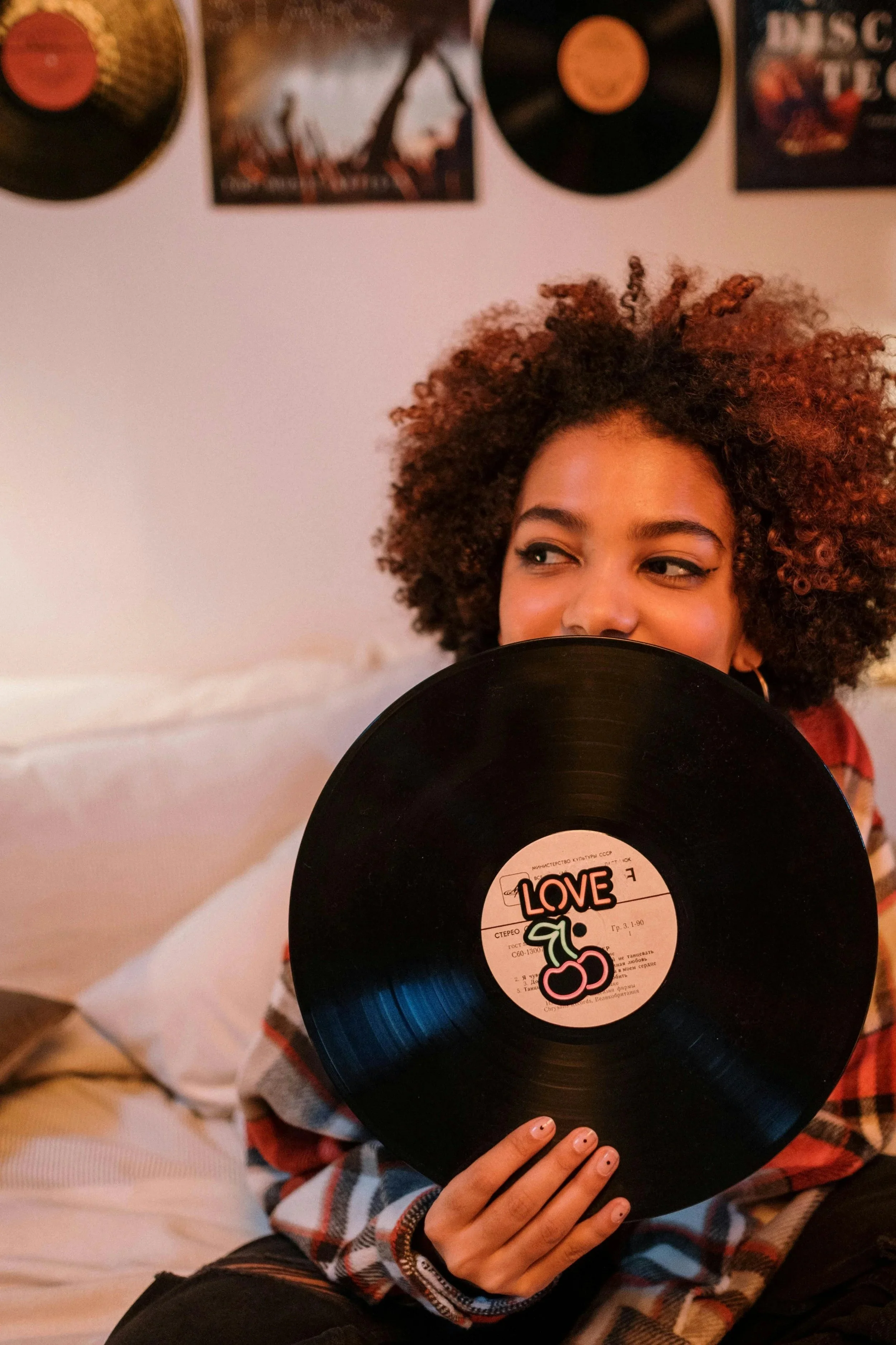 A woman with curly hair holds a vinyl record labeled 'LOVE' with a cherry illustration in front of her face, sitting on a bed with records and posters on the wall behind her.