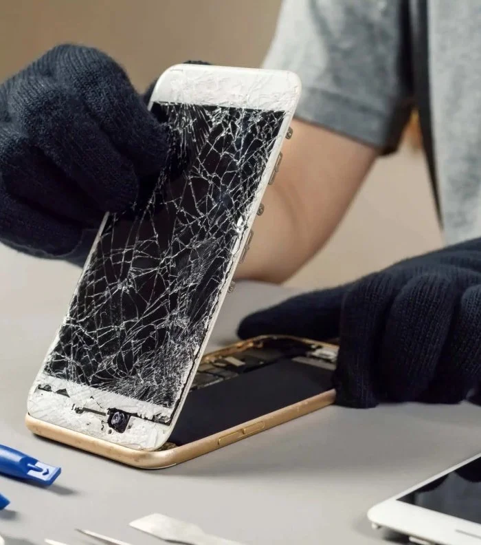 A person wearing black gloves handling a severely broken white smartphone with a shattered screen, on a gray work surface in a repair setting.